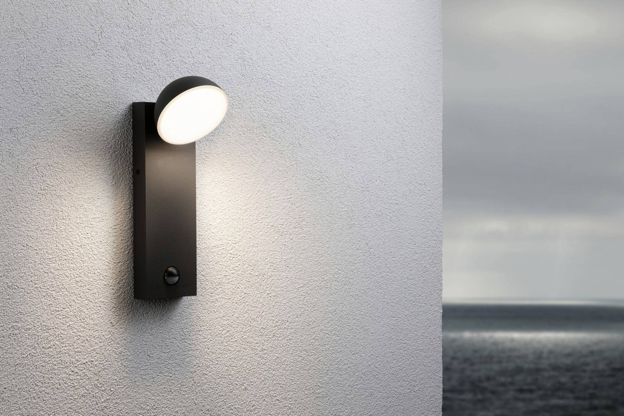 Lampa zewnętrzna na białej ścianie. Światło delikatnie skierowane w dół. W tle widoczne są chmury nad morzem.