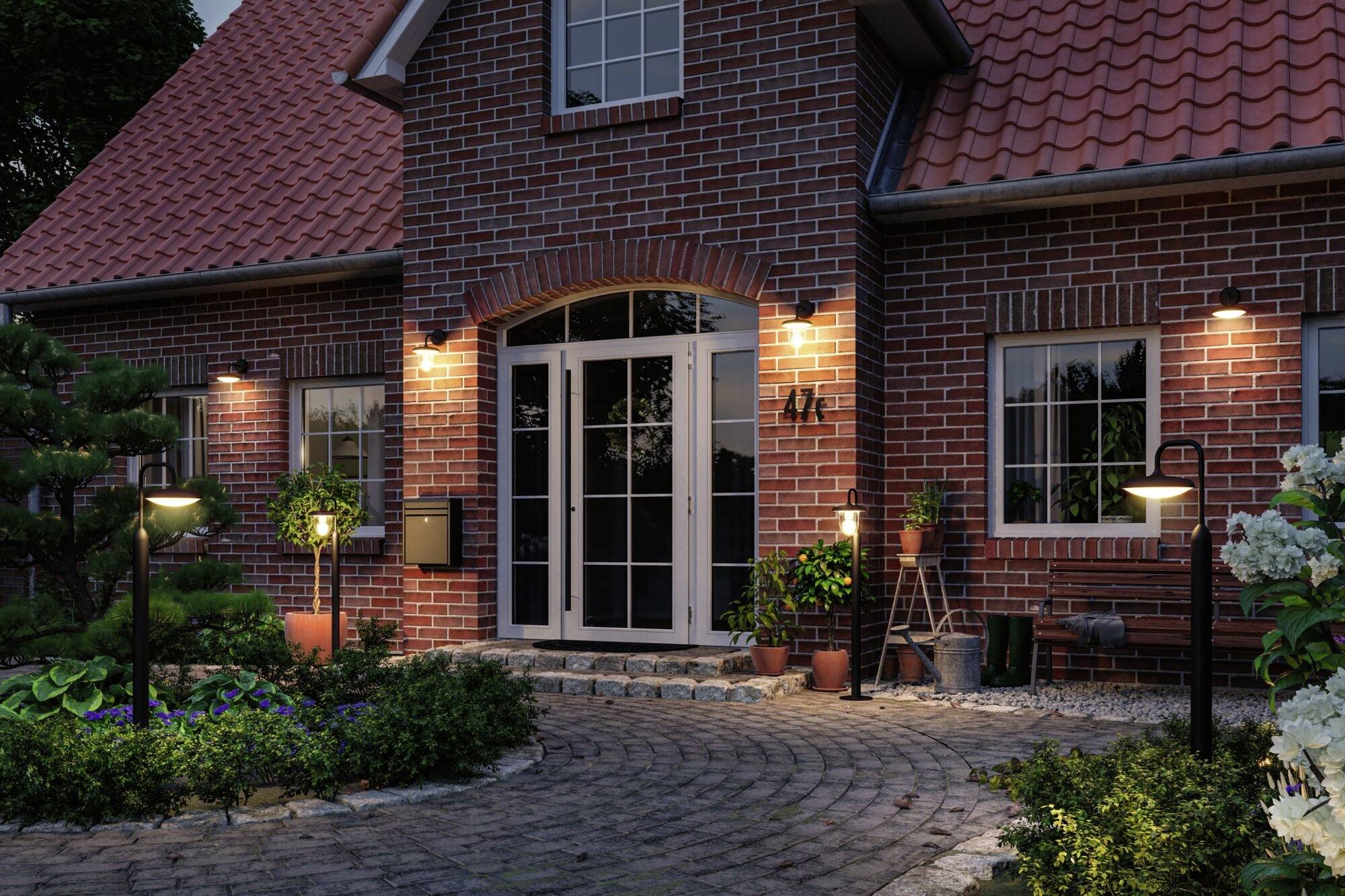 Ceglany dom jednorodzinny z podświetlonym wejściem nocą, otoczony zadbnym ogrodem. Ścieżka z kamiennej kostki prowadzi do drzwi wejściowych.