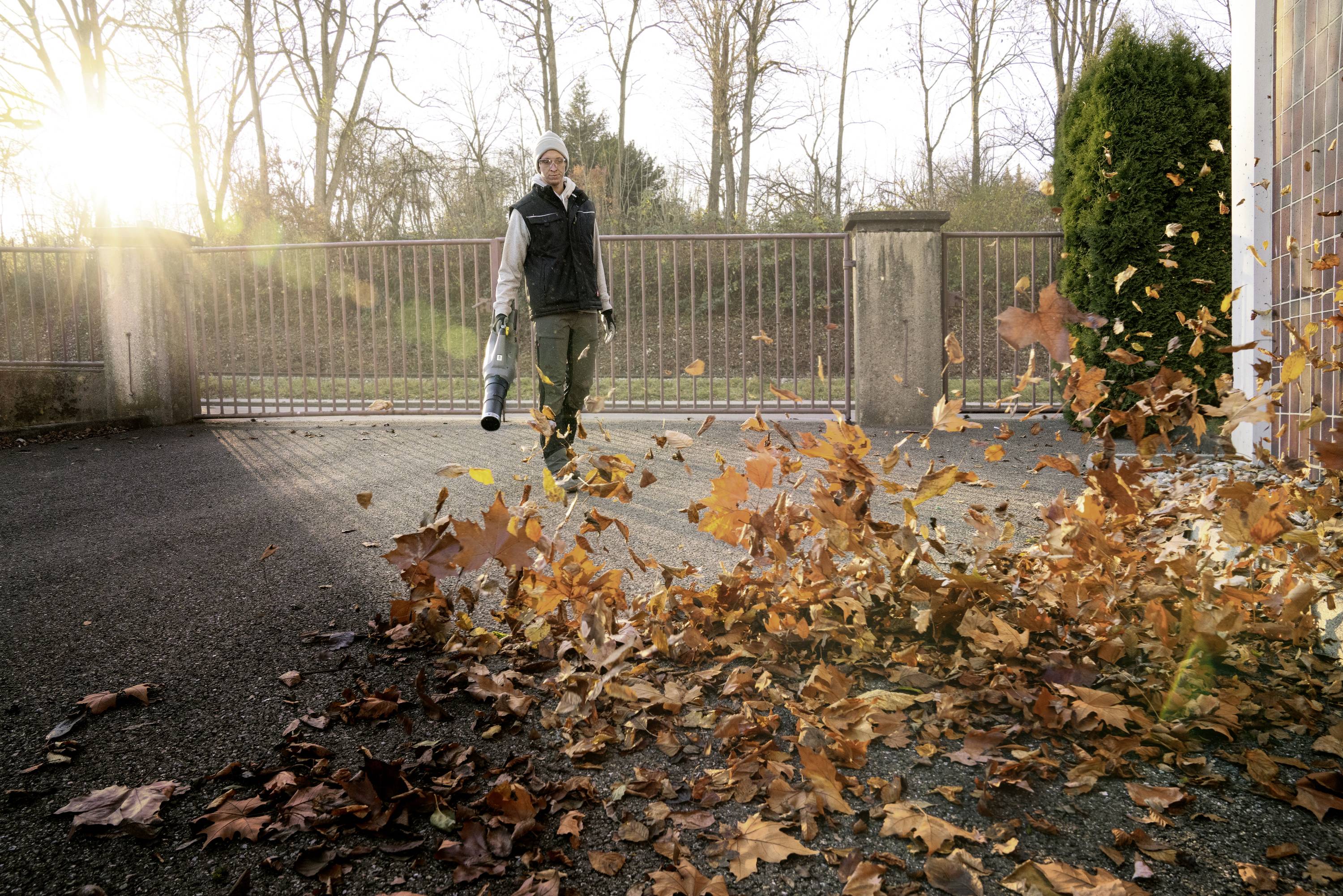 Osoba zdmuchuje jesienne liście dmuchawą na podjeździe, w tle słoneczne promienie.