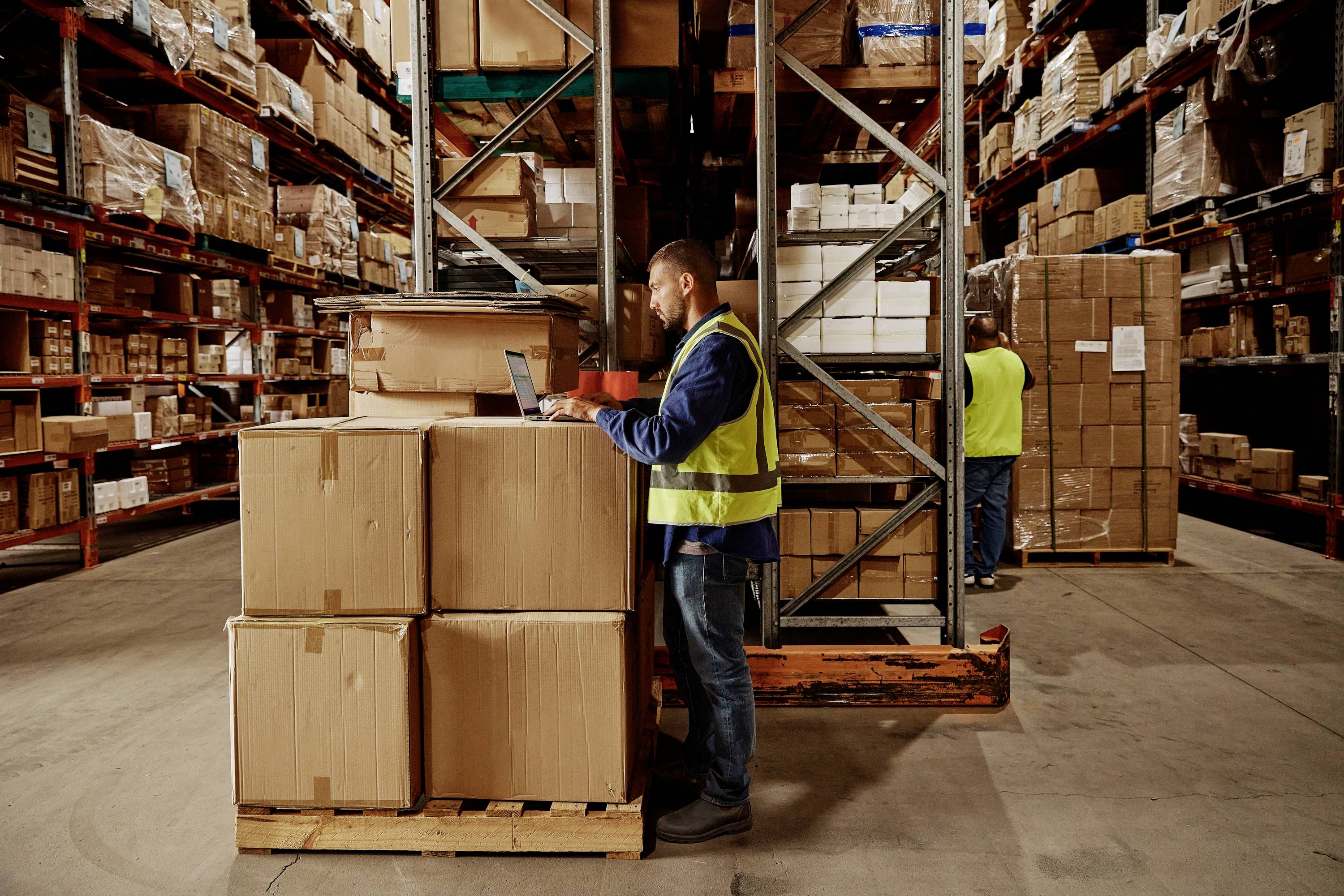Mężczyzna w żółtej kamizelce pracuje na laptopie w magazynie pełnym kartonów i regałów. W tle widać kolejnego pracownika.