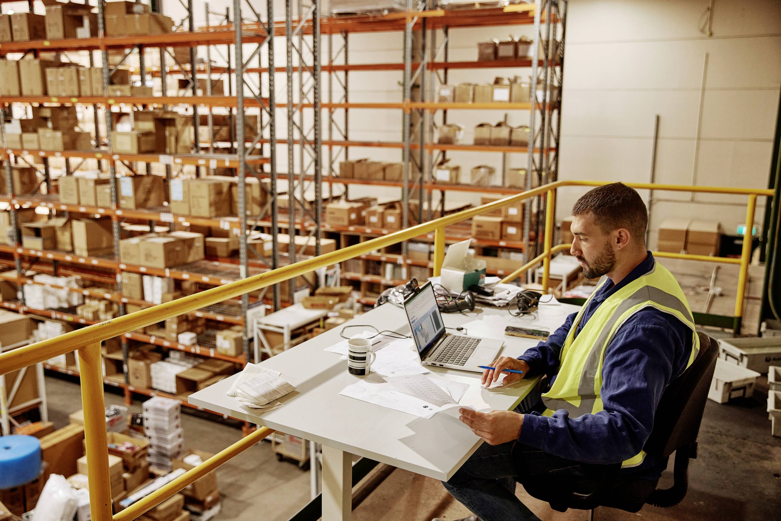 Pracownik w kamizelce odblaskowej pracuje przy biurku w magazynie. W tle widoczne są regały z kartonami.