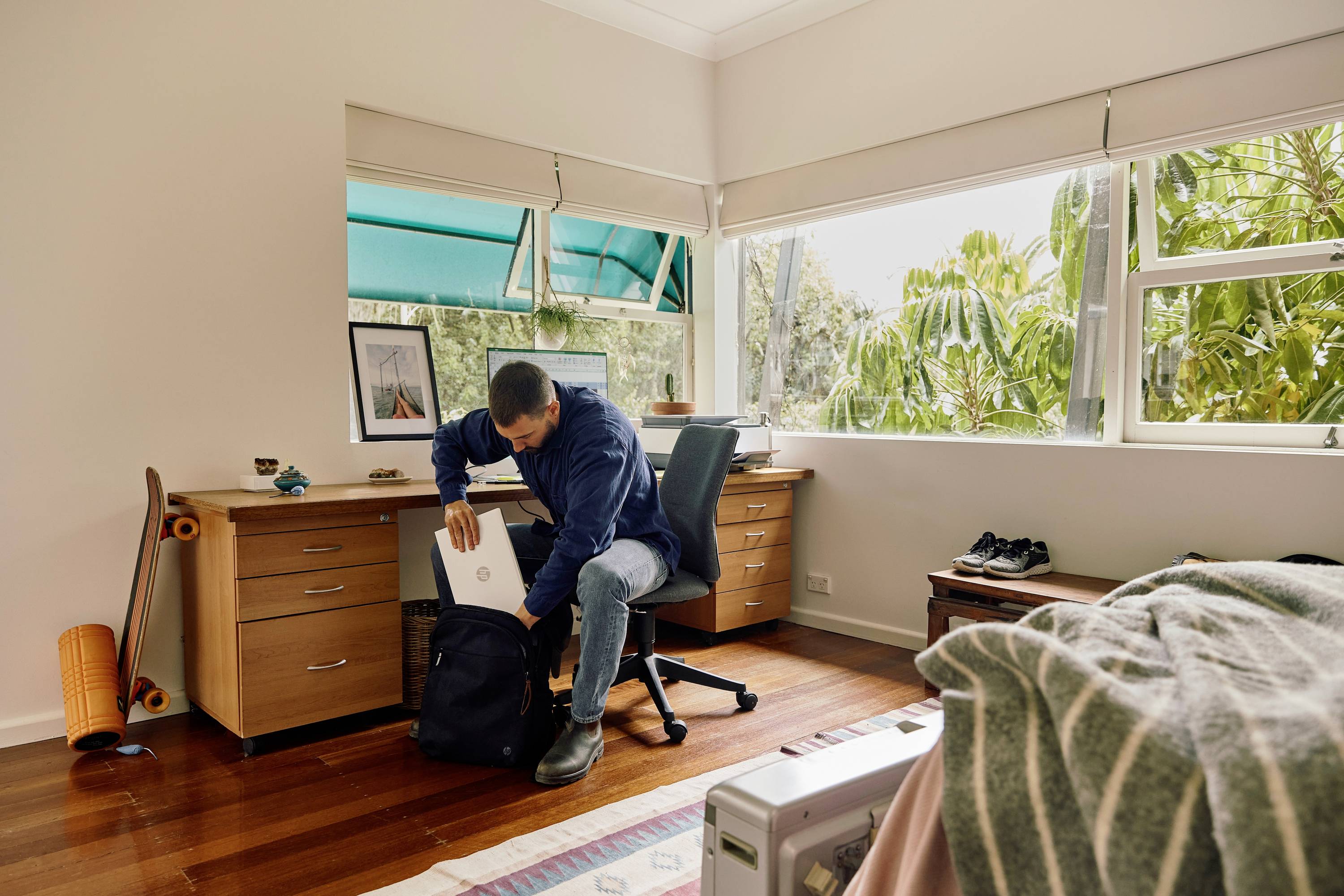 Mężczyzna pakuje laptopa do plecaka w jasnym pokoju z biurkiem, krzesłem, deskorolką i dużymi oknami.