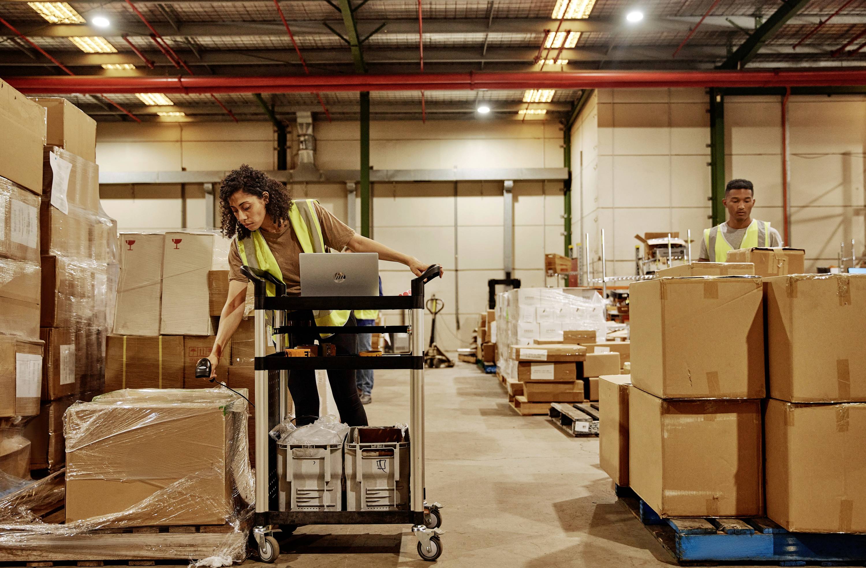 Kobieta w magazynie skanuje pudło za pomocą ręcznego skanera. W tle mężczyzna transportuje kolejne paczki.