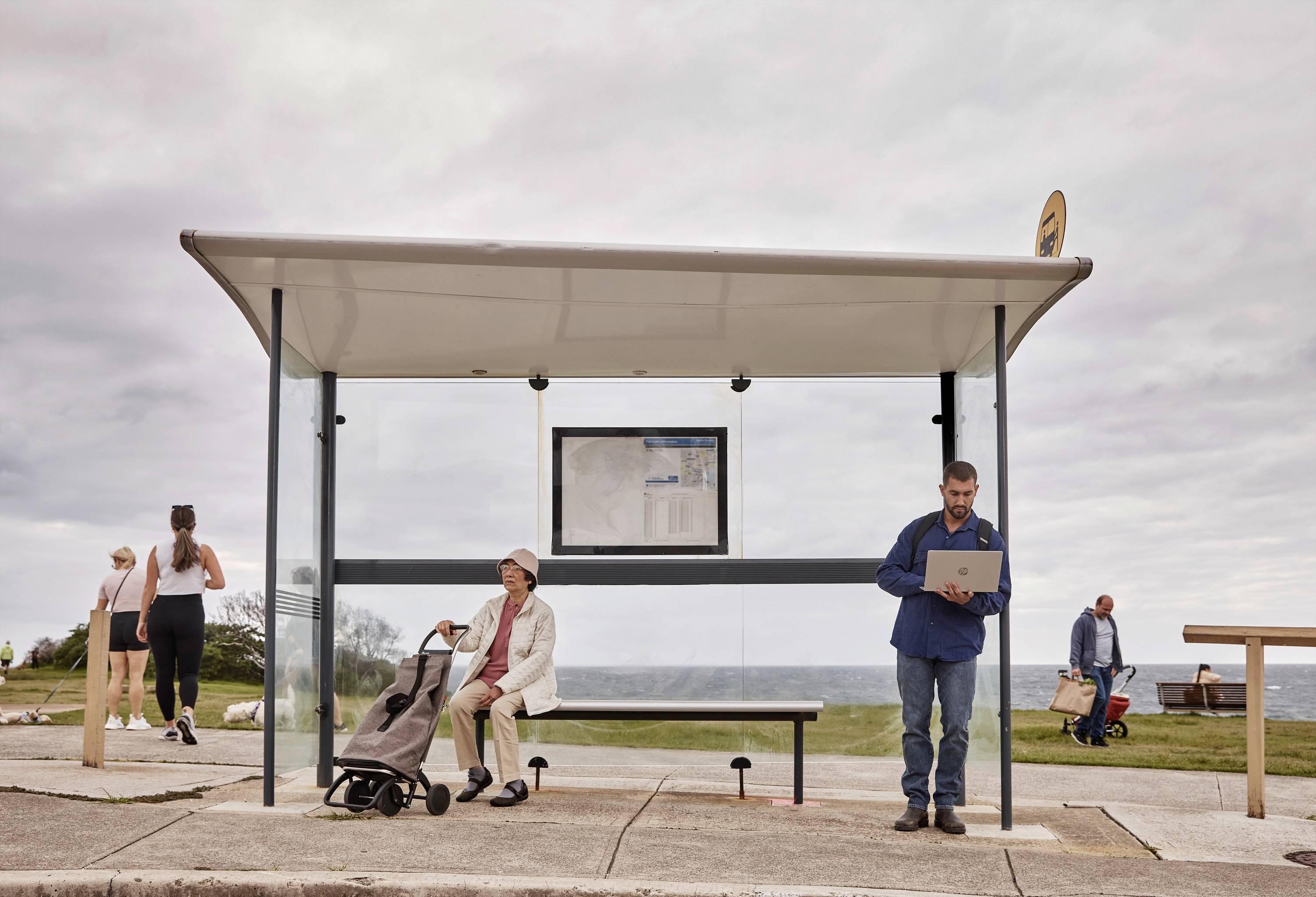 Przystanek autobusowy na plaży, gdzie siedzi starszy mężczyzna i stoi drugi mężczyzna z laptopem. W tle spacerujący ludzie na tle pochmurnego nieba.