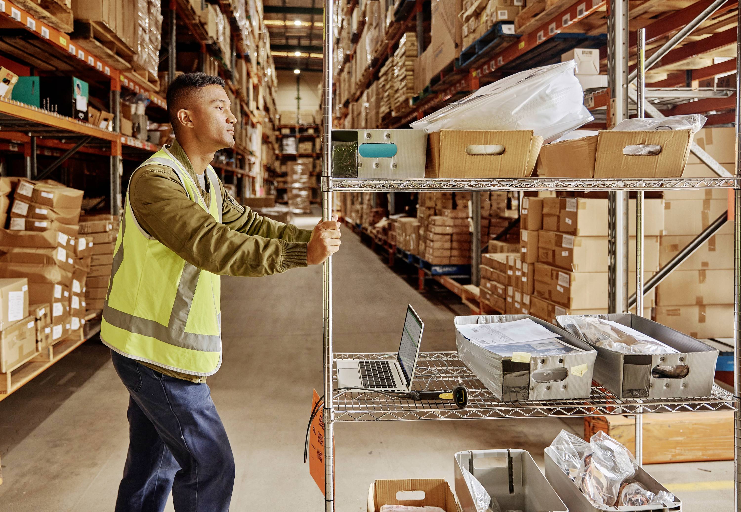 Osoba w magazynie przesuwa regał na kółkach. W tle widoczne są regały z kartonami. Na jednym z regałów leży laptop.