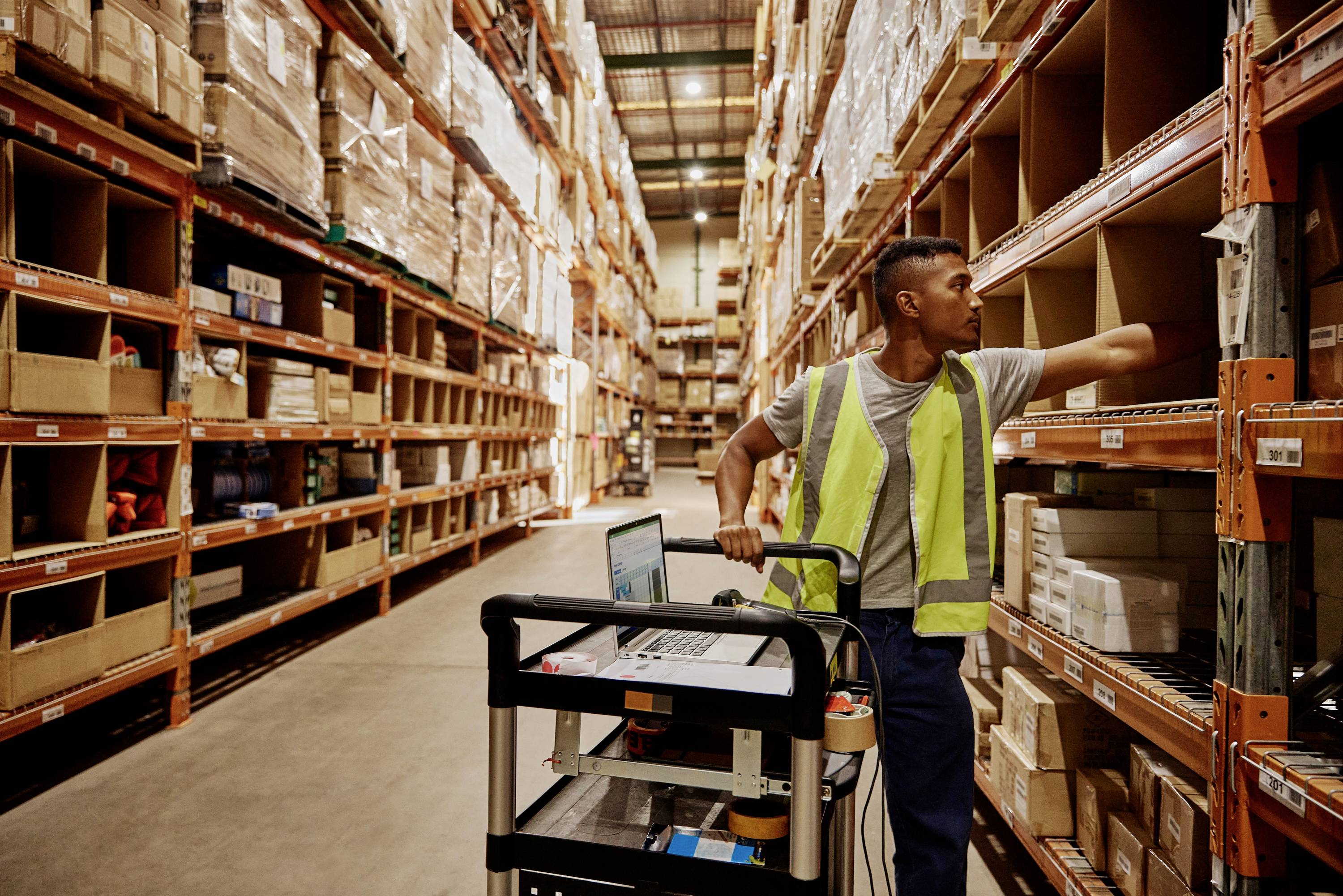 Osoba w żółtej kamizelce odblaskowej pracuje w magazynie, obsługuje laptopa na wózku i sięga po towary z regału.