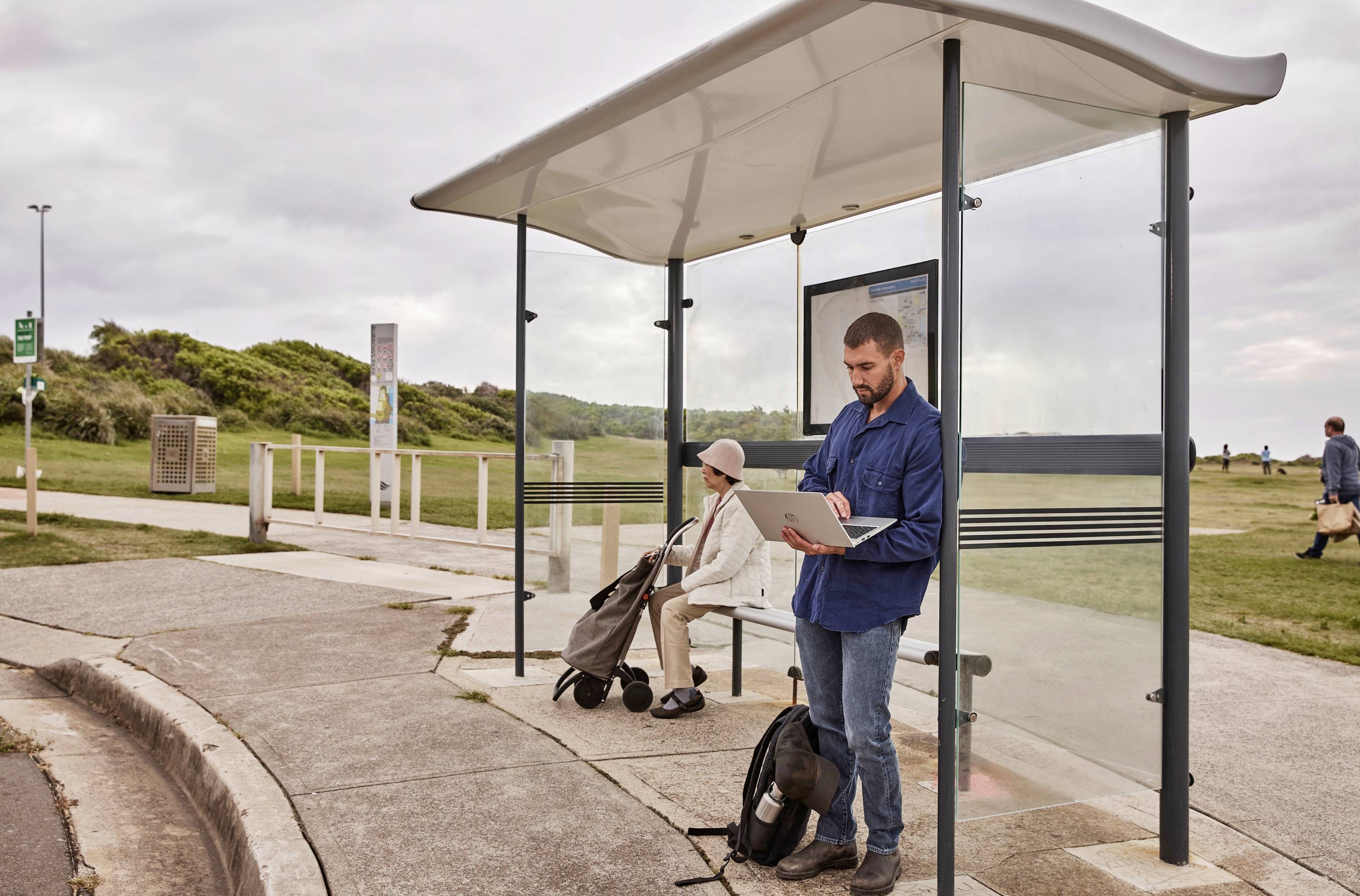Mężczyzna pracuje na laptopie na przystanku autobusowym, podczas gdy kobieta siedzi i czeka. W tle widać park.