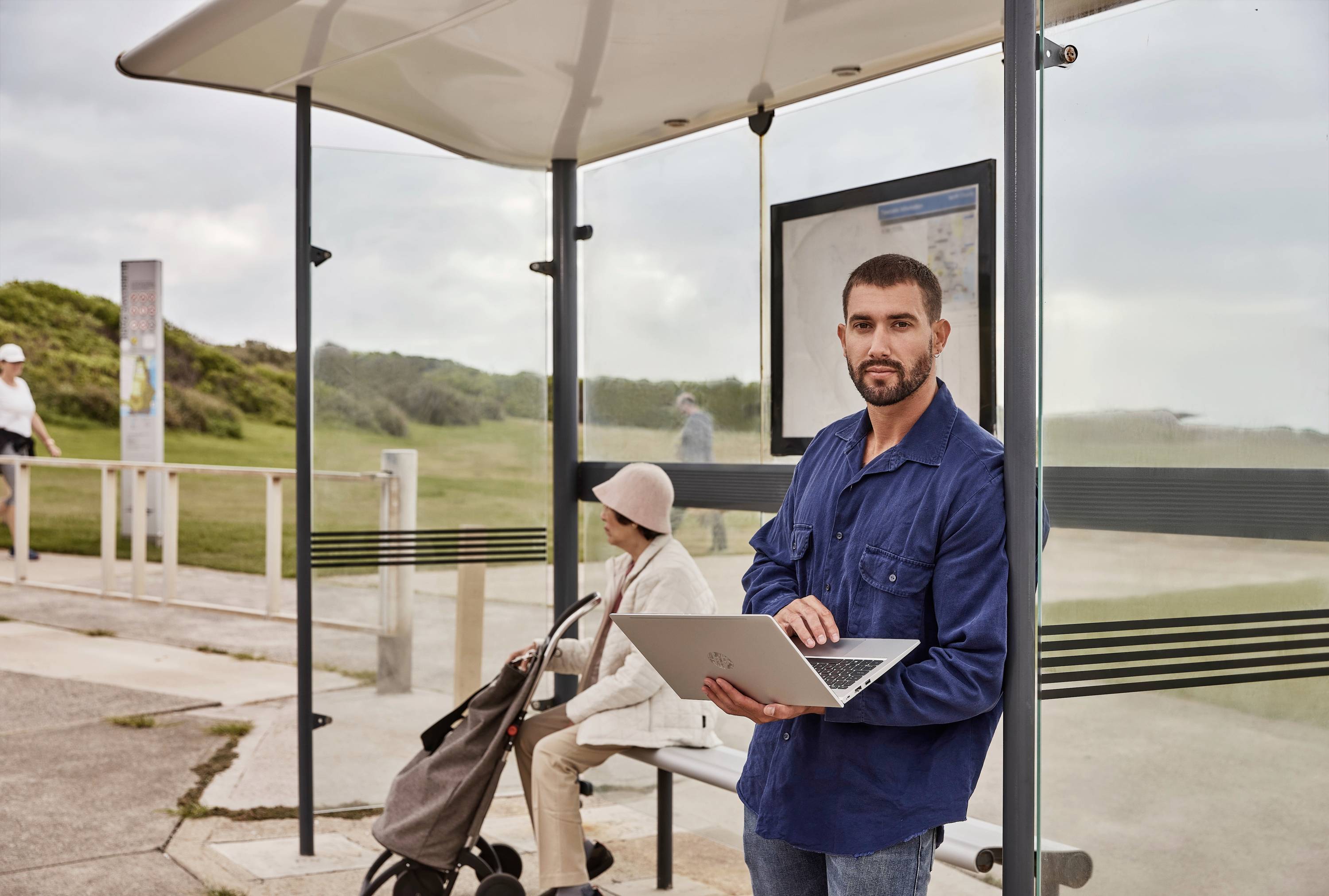 Mężczyzna stoi na przystanku autobusowym z laptopem w ręku, a w tle starsza osoba siedzi na ławce i czeka.