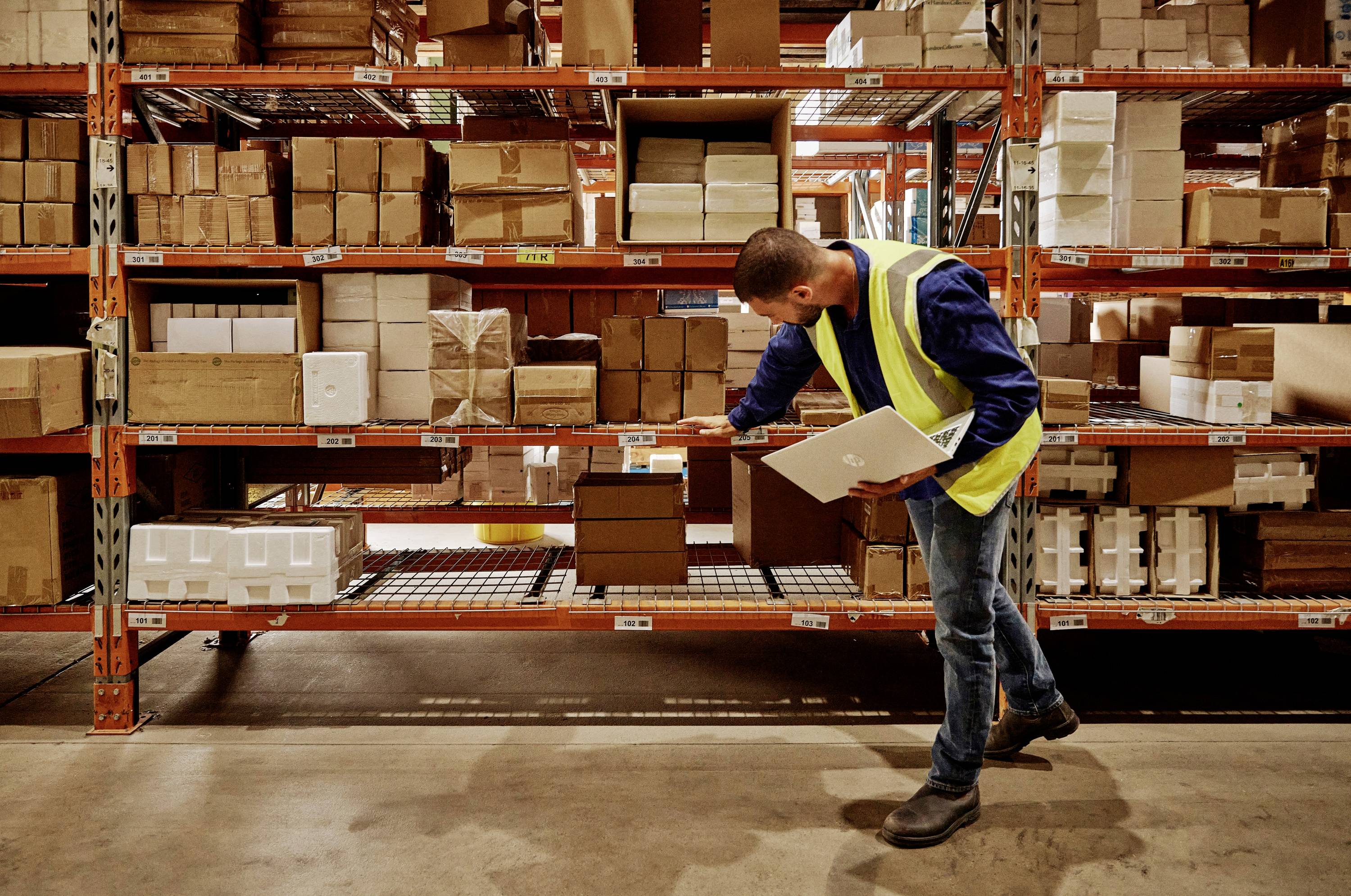 Pracownik magazynu w żółtej kamizelce ochronnej sprawdza stan zapasów na regale pełnym kartonów, trzymając w ręku schowek.
