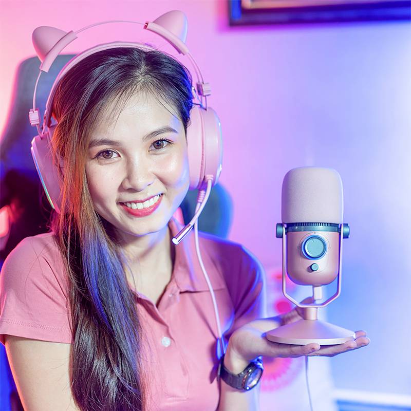 Uśmiechnięta osoba siedzi przed mikrofonem, nosząc słuchawki w kształcie kocie uszy. Ma na sobie różową koszulkę polo.