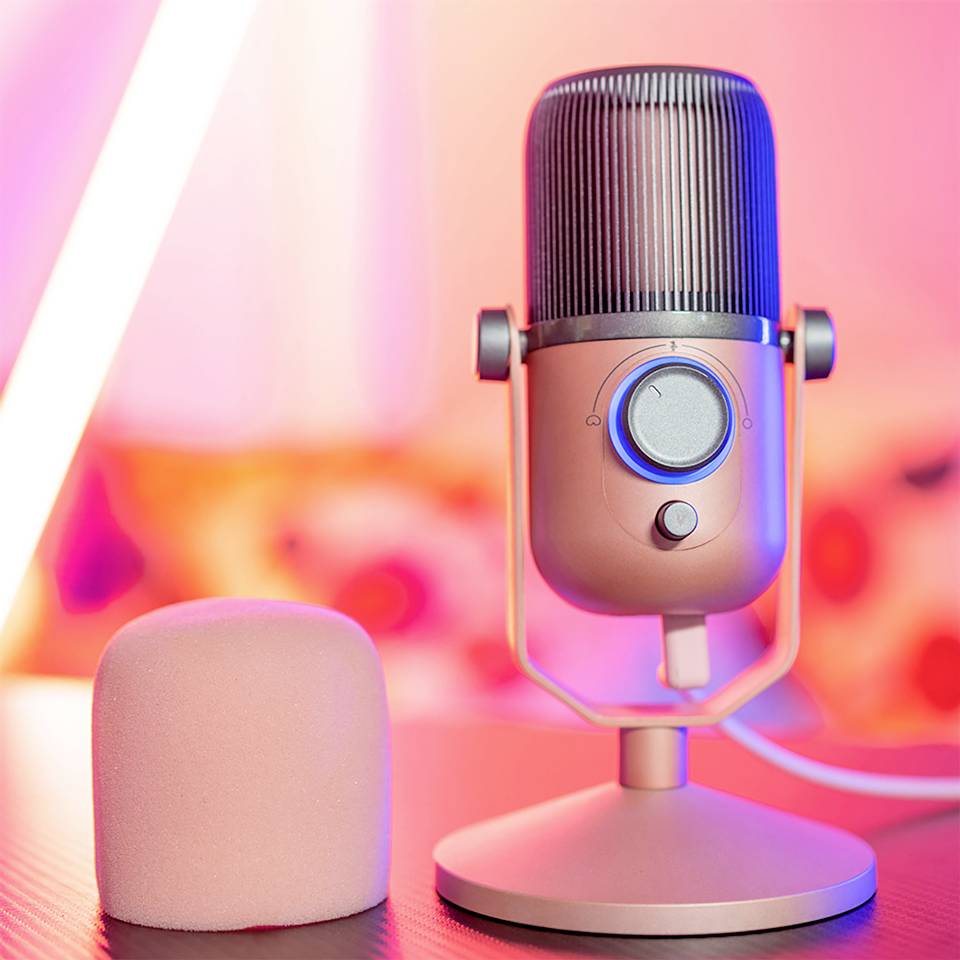 Różowy mikrofon na stole na tle rozmytego, ciepłego tła. Po lewej stronie obok niego okrągła, różowa osłona z pianki.