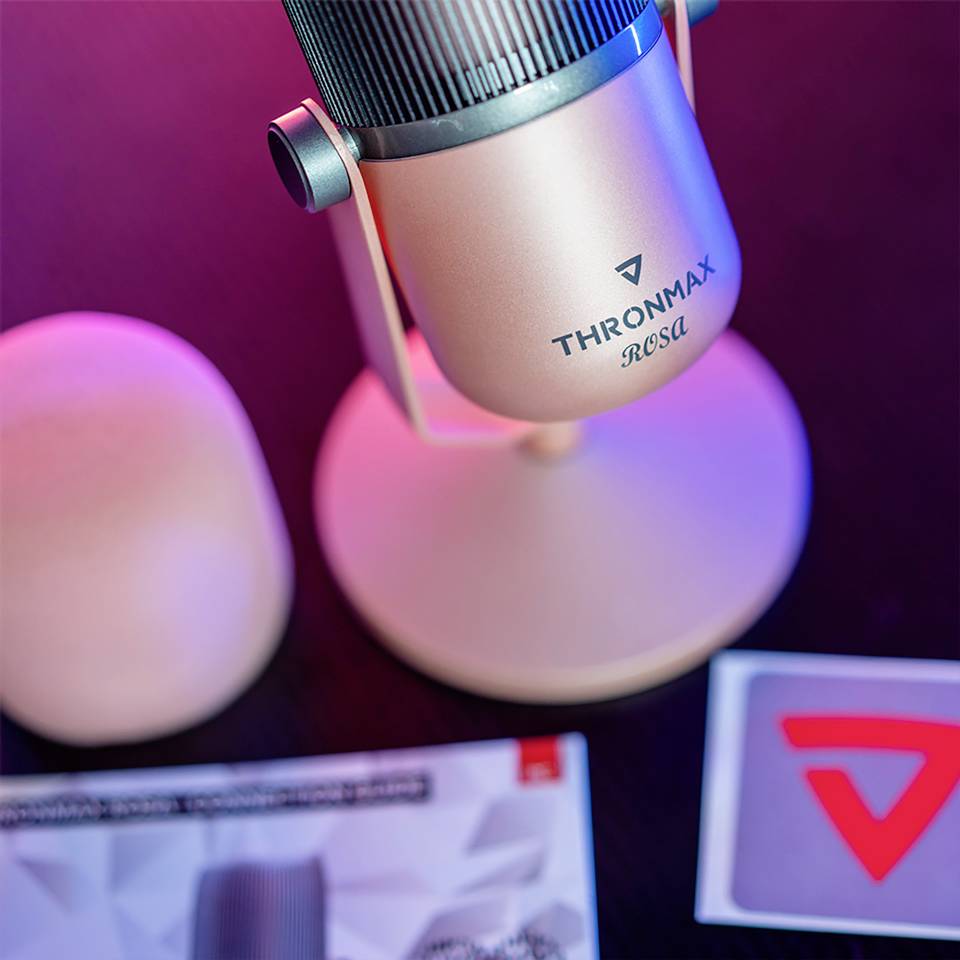 Różowy mikrofon z napisem 'Thronmax Rosa' stoi na stole. Obok niego znajduje się okrągły przedmiot i zeszyt z logo.