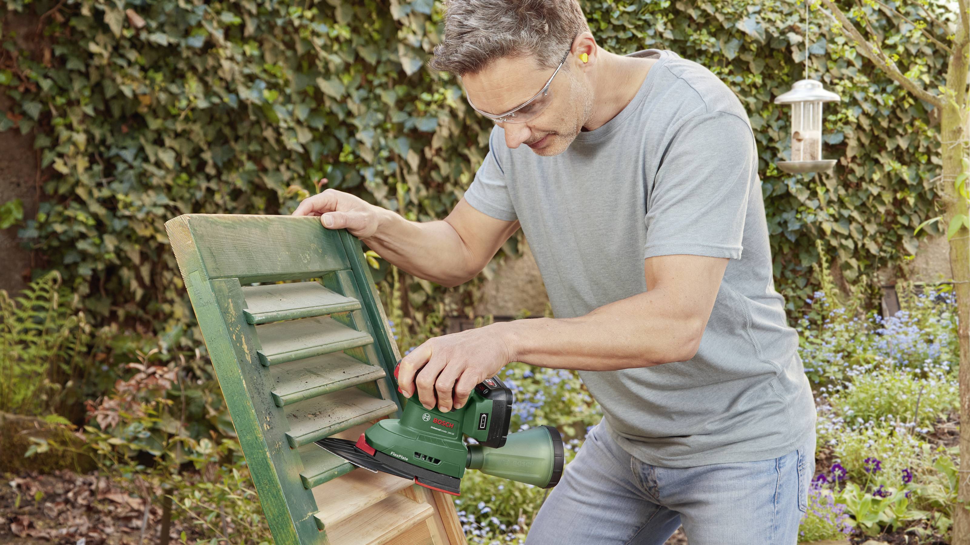 Mężczyzna szlifuje zielone okiennice elektryczną szlifierką w ogrodzie. W tle: bluszcz i domek dla ptaków.