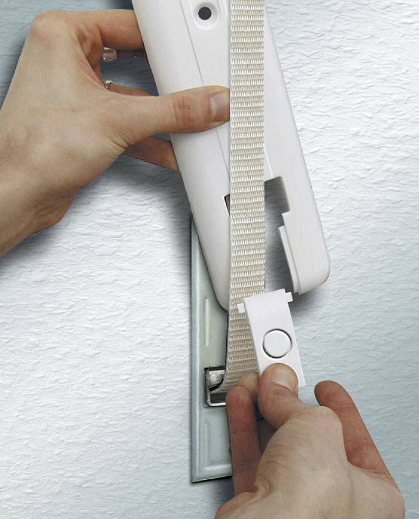 Ręce trzymają nawijacz paska przy ścianie, obsługując system rolet. Pas prowadzony jest przez białe urządzenie.