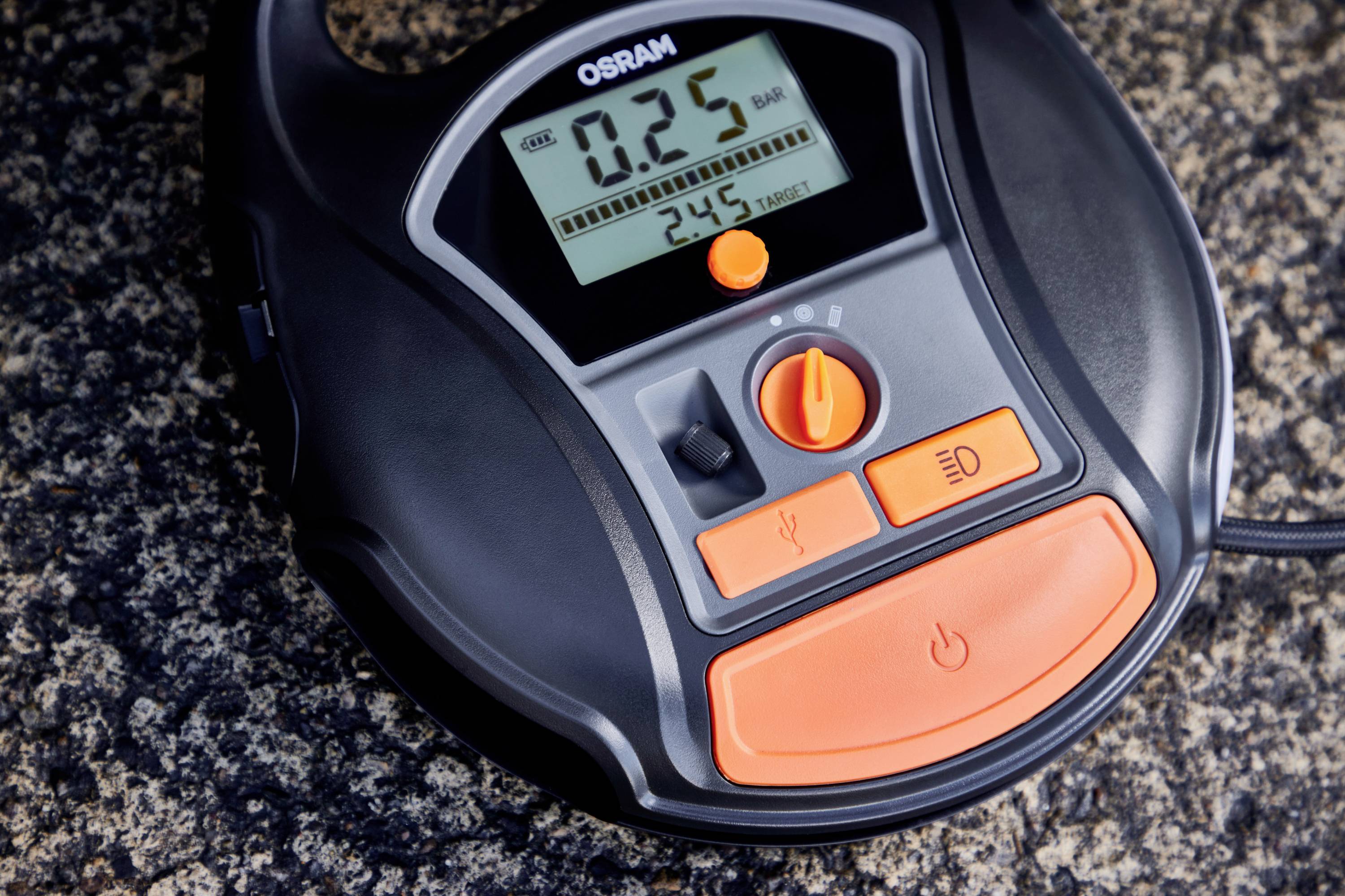 Cyfrowe urządzenie z wyświetlaczem pokazuje '0,25 BAR'. Posiada elementy sterujące do pomiaru ciśnienia powietrza oraz dwa pomarańczowe przyciski.