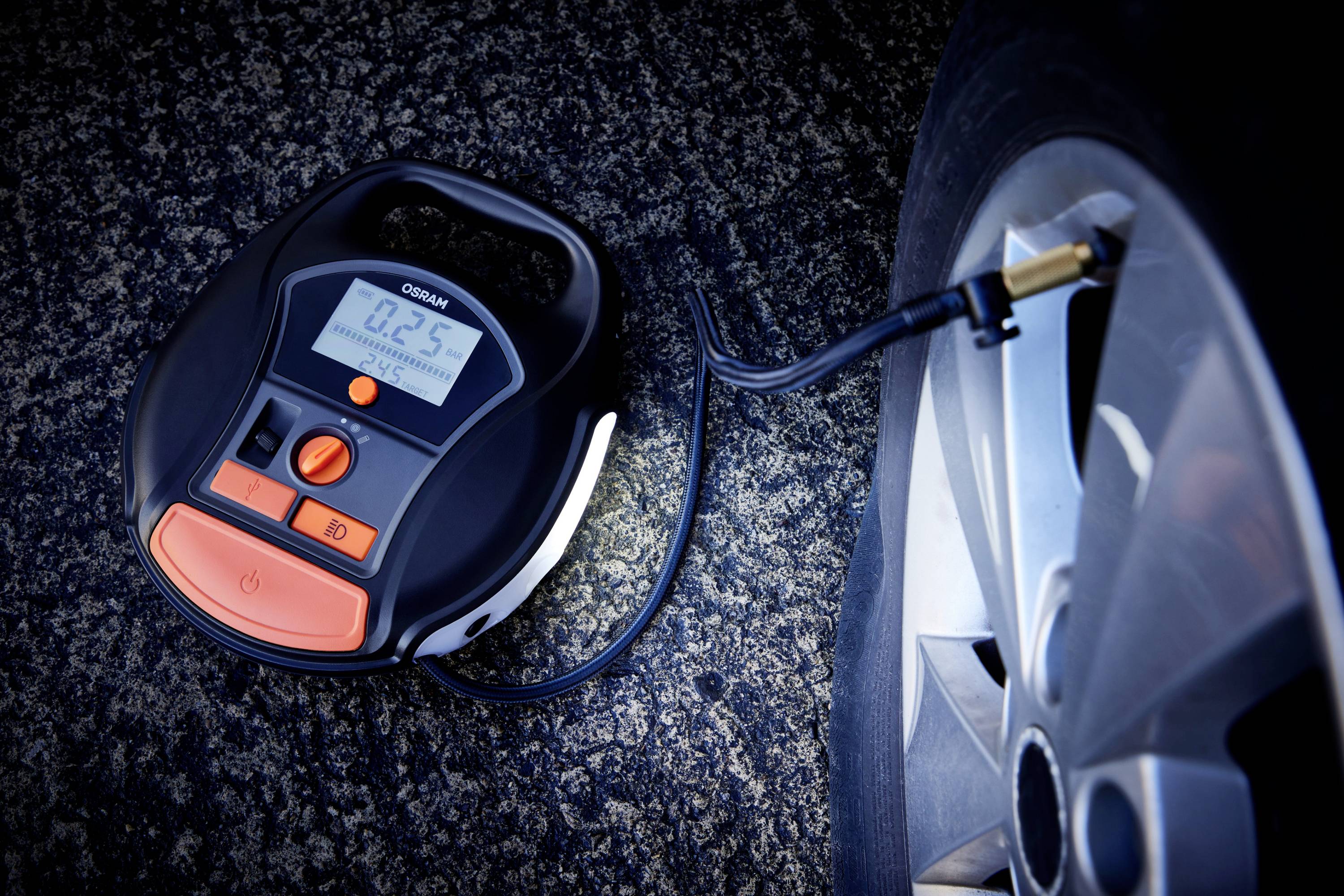 Cyfrowa pompka sprężająca napełnia oponę samochodu. Ekran wyświetla '0,25' bara. W tle widać drogę.
