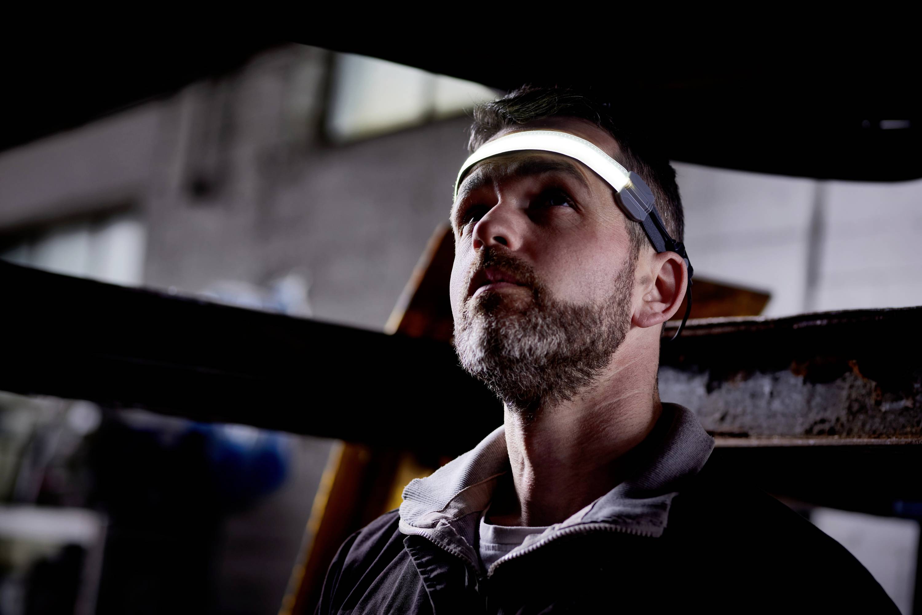 Mężczyzna z brodą nosi świecącą opaskę na głowie. Spogląda w górę, jakby coś kontrolował lub obserwował.