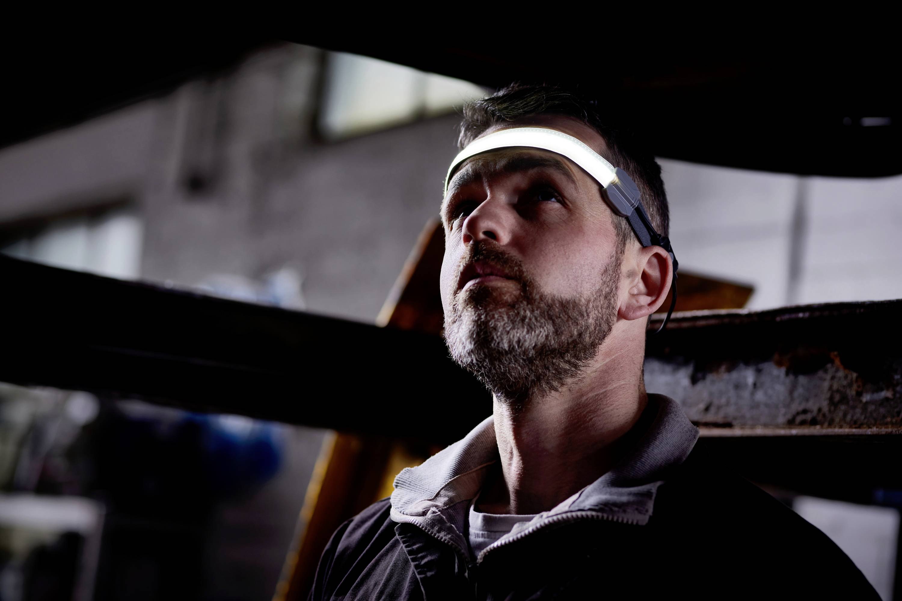 Mężczyzna nosi świecącą opaskę na czole w ciemnym pomieszczeniu, patrzy w górę, otoczony stalowymi belkami.