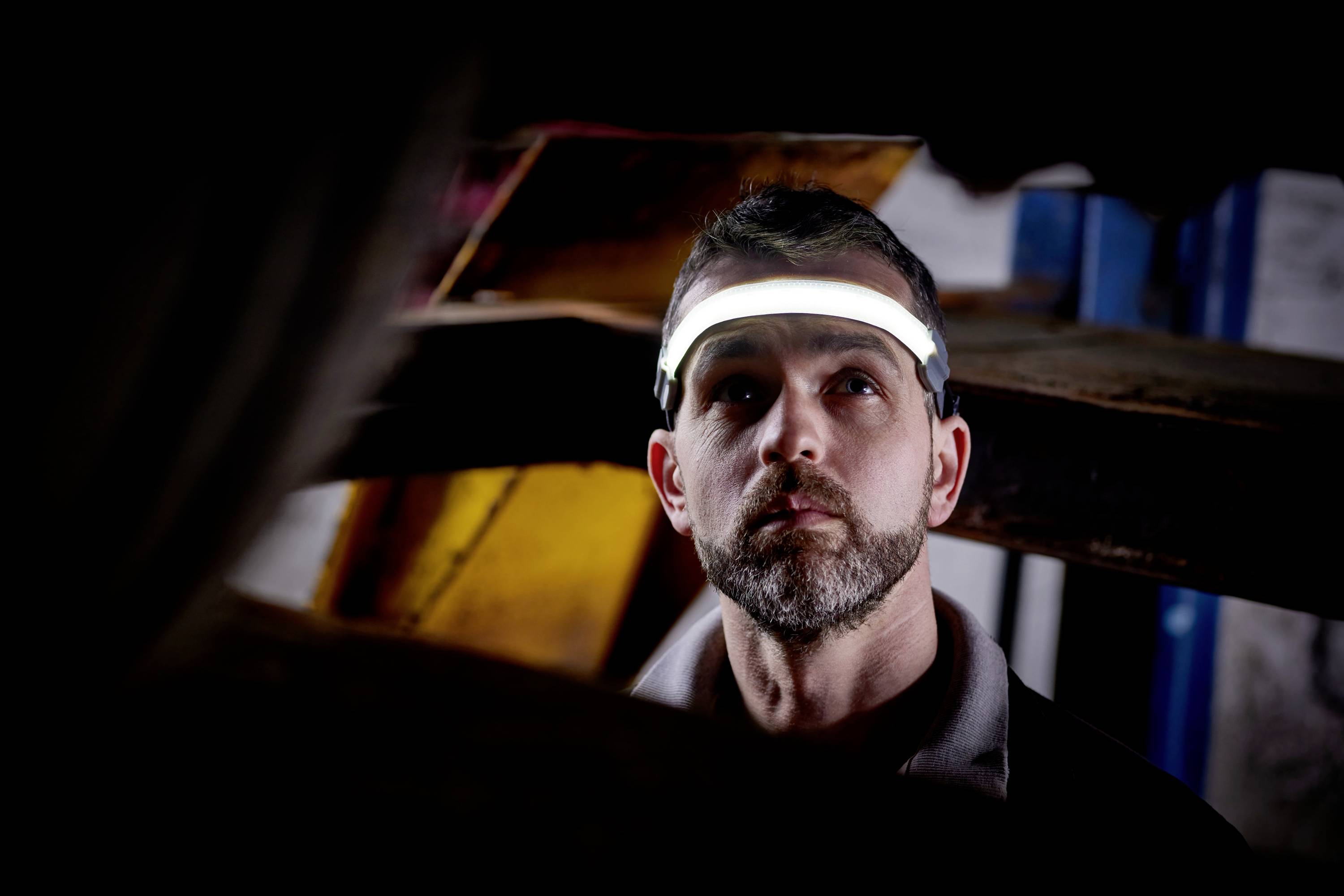 Mężczyzna z brodą nosi świecącą opaskę na głowie i patrzy w górę, jakby coś badał. W tle widoczne są drewniane półki.