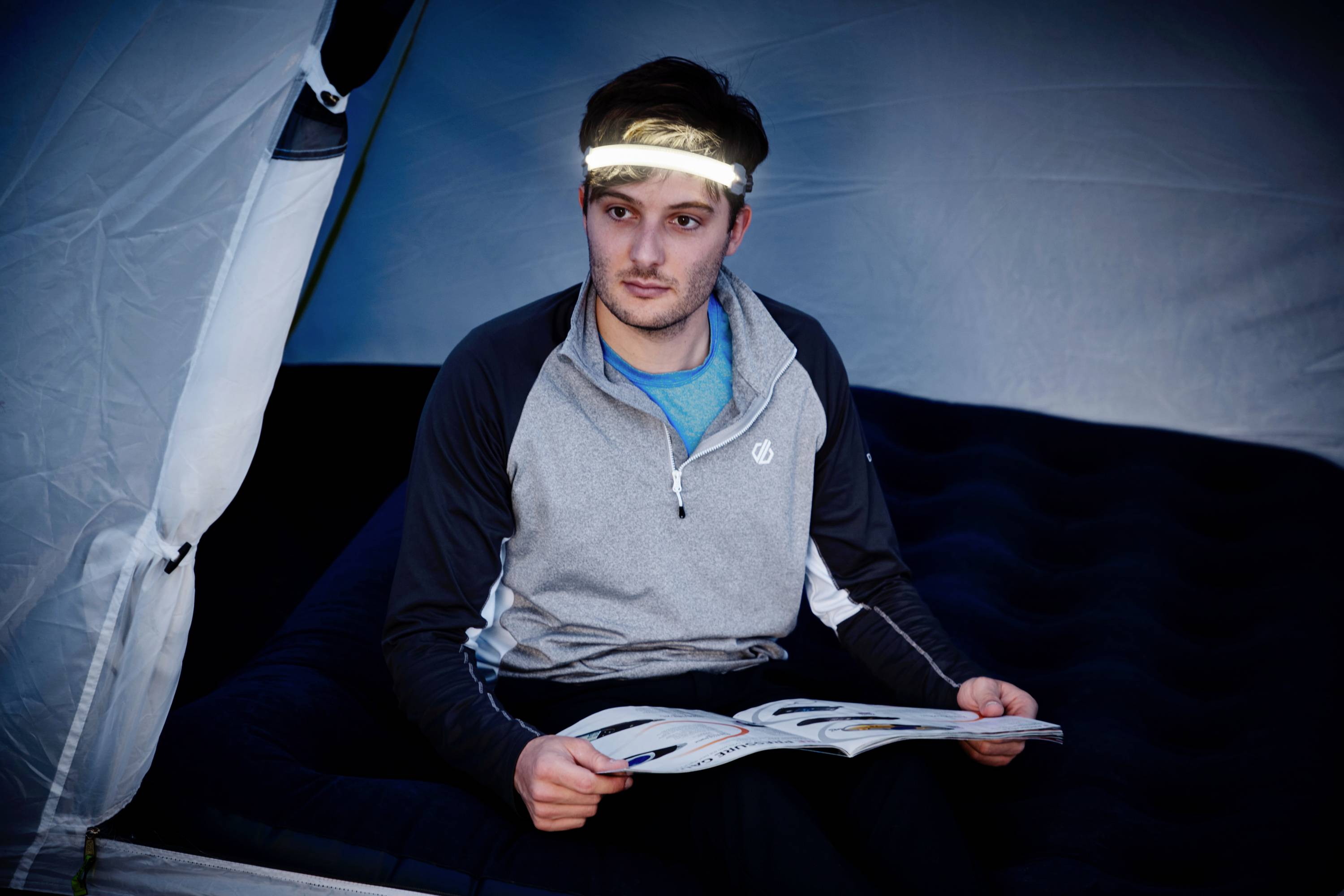 Jedna osoba siedzi w namiocie na materacu powietrznym i czyta magazyn. Ma na głowie opaskę z lampką, która oświetla jej twarz.
