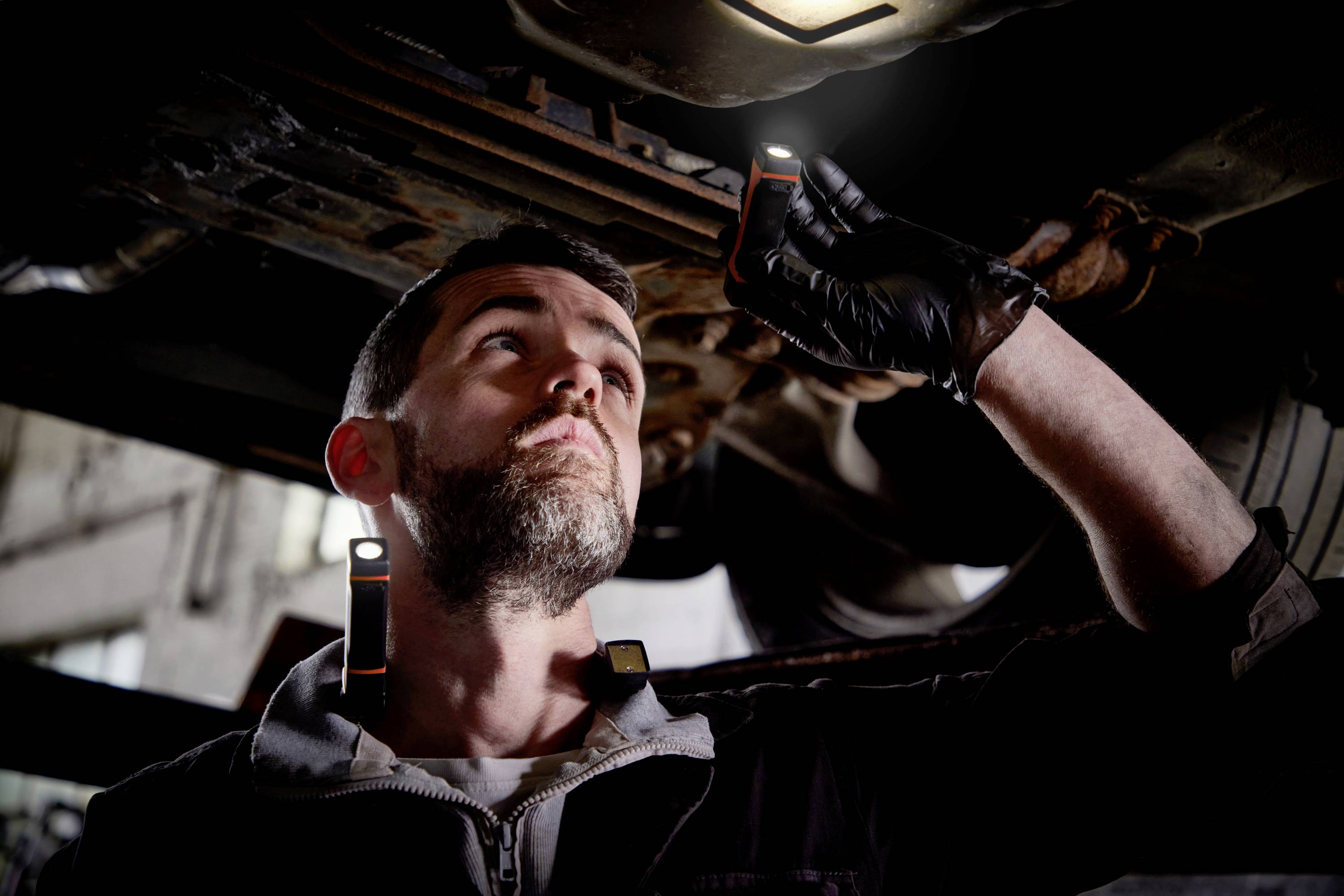 Mechanik sprawdza pod pojazdem za pomocą latarki. Ma na sobie czarną kurtkę i czarne rękawice.