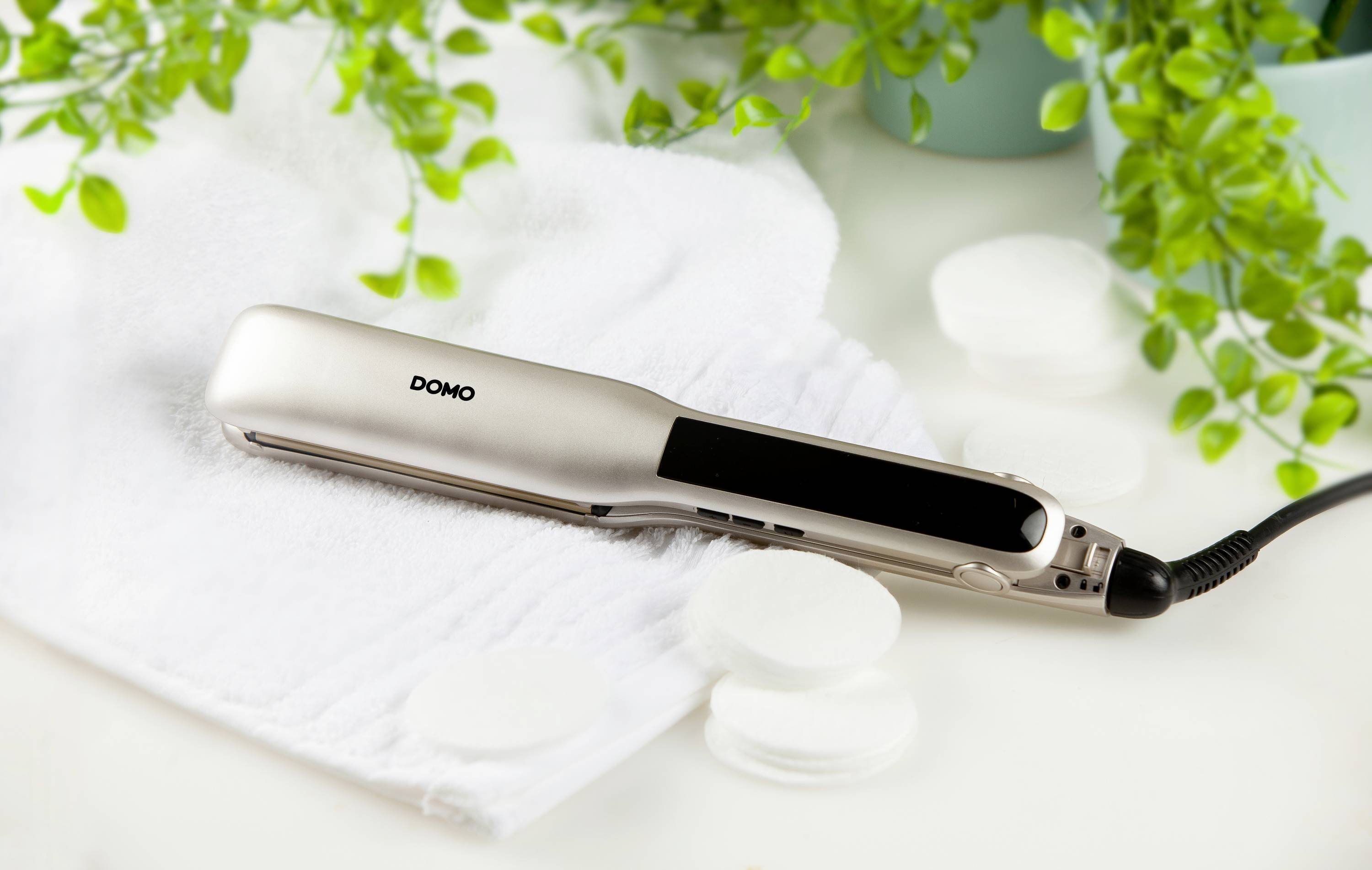 Prostownica Domo leży na ręczniku, otoczona roślinami i wacikanymi płatkami.