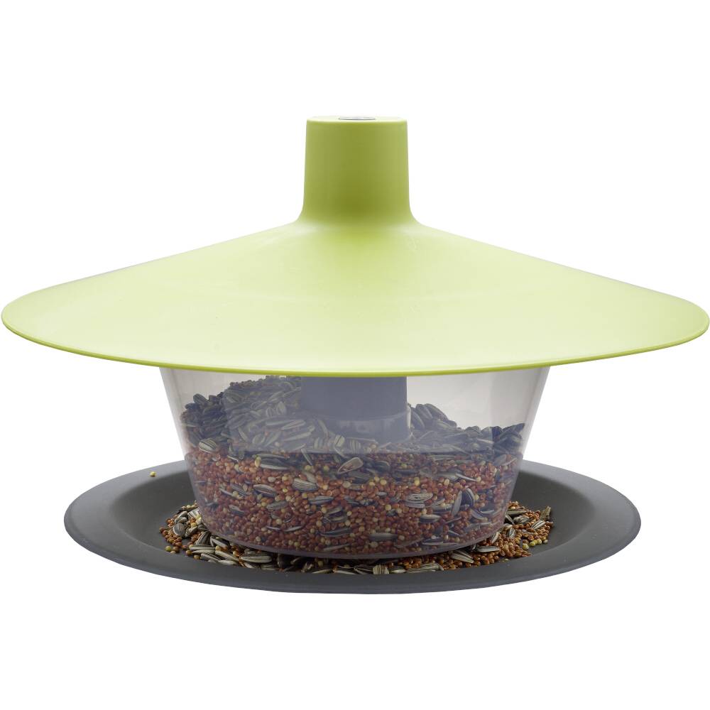 Zielony domek dla ptaków z daszkiem w kształcie pokrywy, wypełniony różnymi nasionami, umieszczony na okrągłym szarym talerzyku.