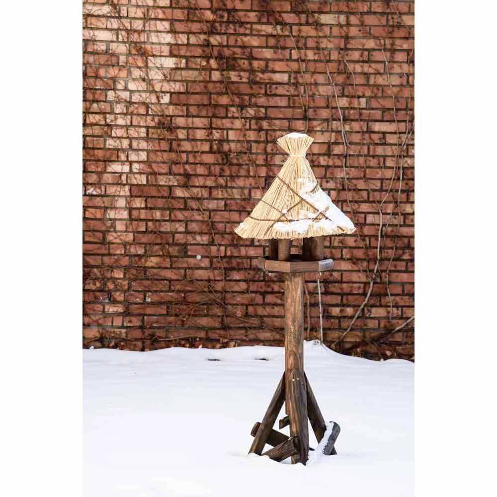 Domek dla ptaków ze słomianym dachem na drewnianej nodze w śnieżnym podwórzu przed ceglastoczerwonym murem.