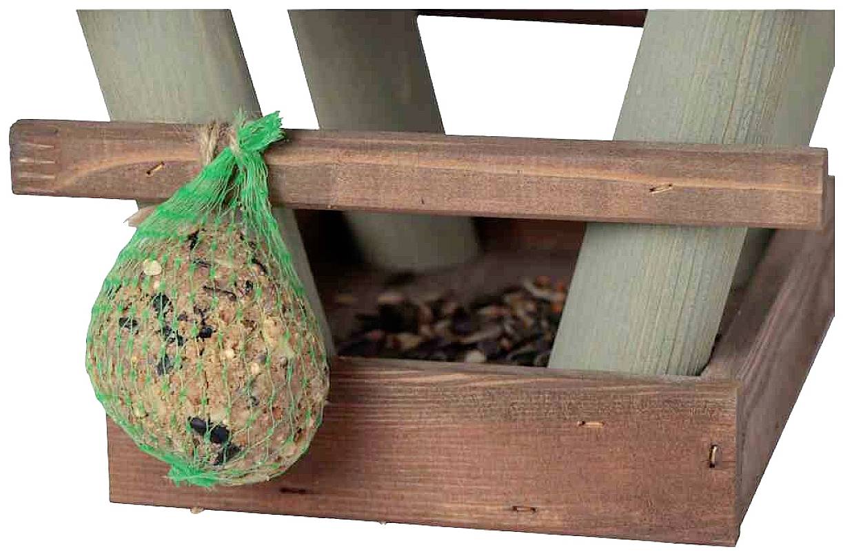 Drewniany podajnik karmy dla ptaków z siatką pełną ziaren wisi z boku. W tle widoczne są drewniane belki.