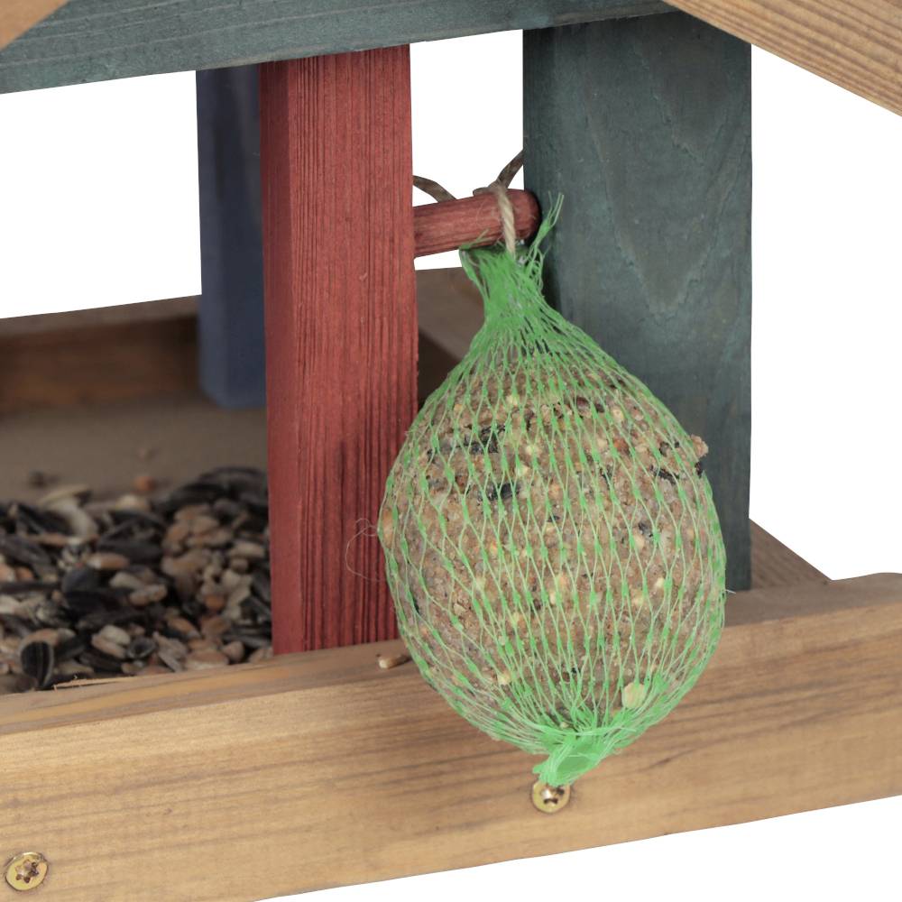 Domek dla ptaków z zielona siatką pełną kul z pokarmem wisi obok. Poniżej rozrzucone są różne nasiona i ziarna.