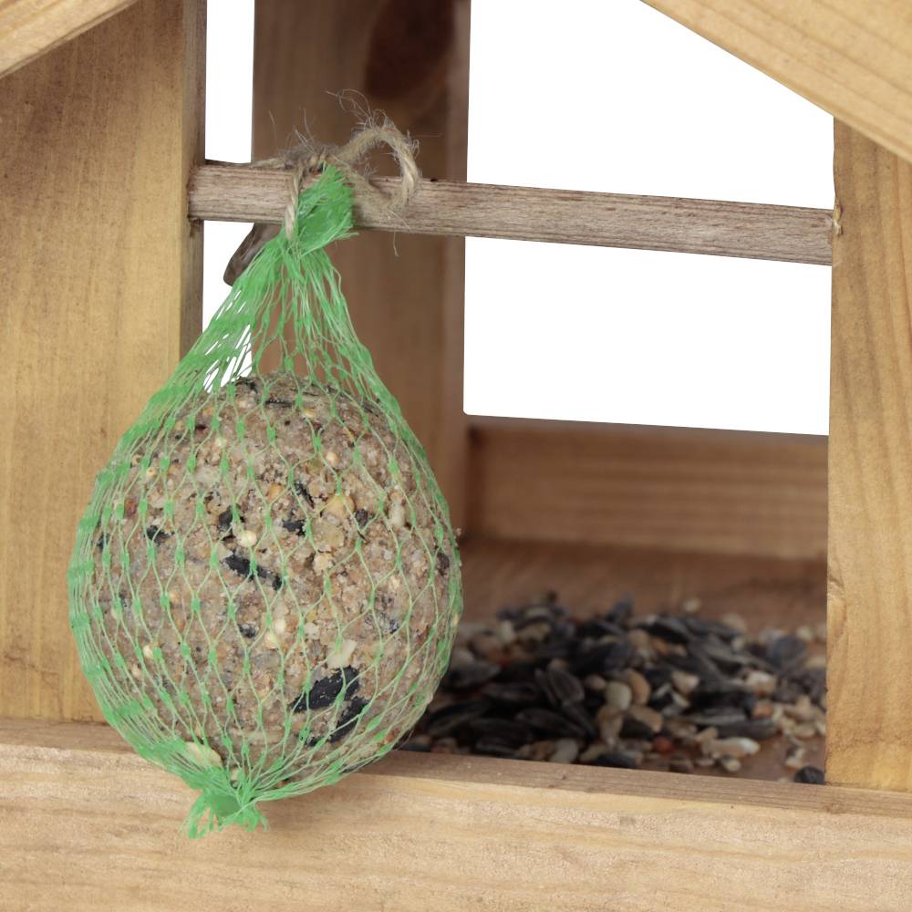Drewniany domek dla ptaków z kulką tłuszczową w zielona siatce. W tle widoczne są luźno rozrzucone ziarna i mieszanki nasienne.