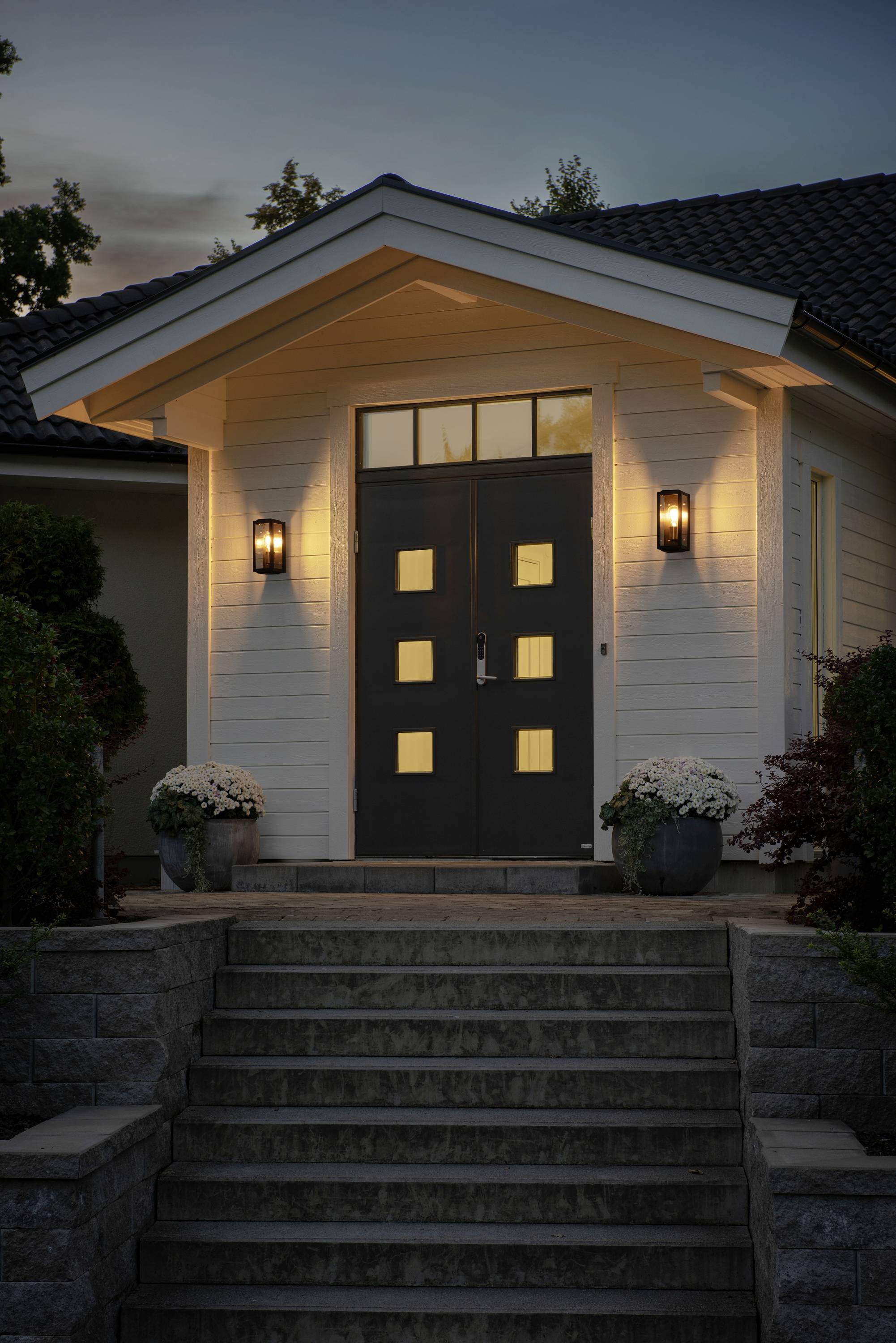Wejście do domu oświetlonego o zmierzchu, z symetrycznymi schodami, doniczkami z roślinami i kwadratowymi oknami przy drzwiach.