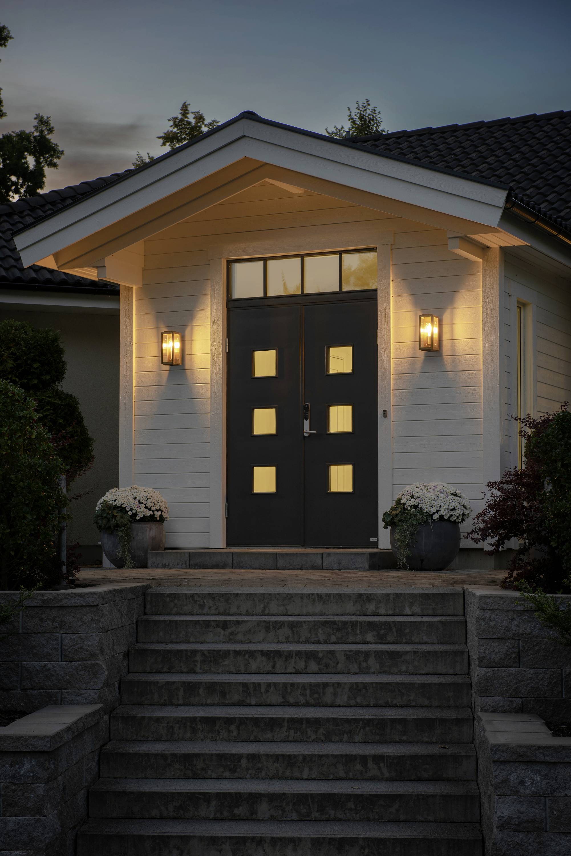 Wejście do domu o zmierzchu z oświetlonymi drzwiami, dwiema lampami bocznymi, schodami i roślinami po obu stronach.