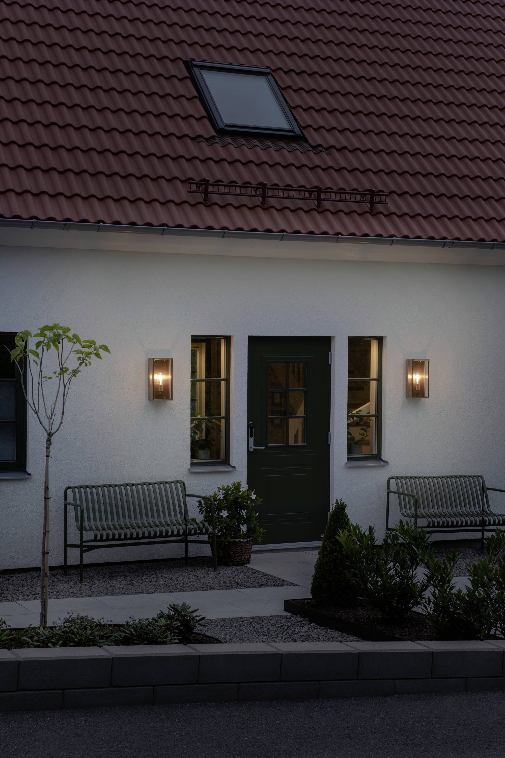 Dom z czerwonym dachem ceramicznym i oknami dachowymi. Przed wejściem stoją dwie ławki i roślina, oświetlone lampami ściennymi.