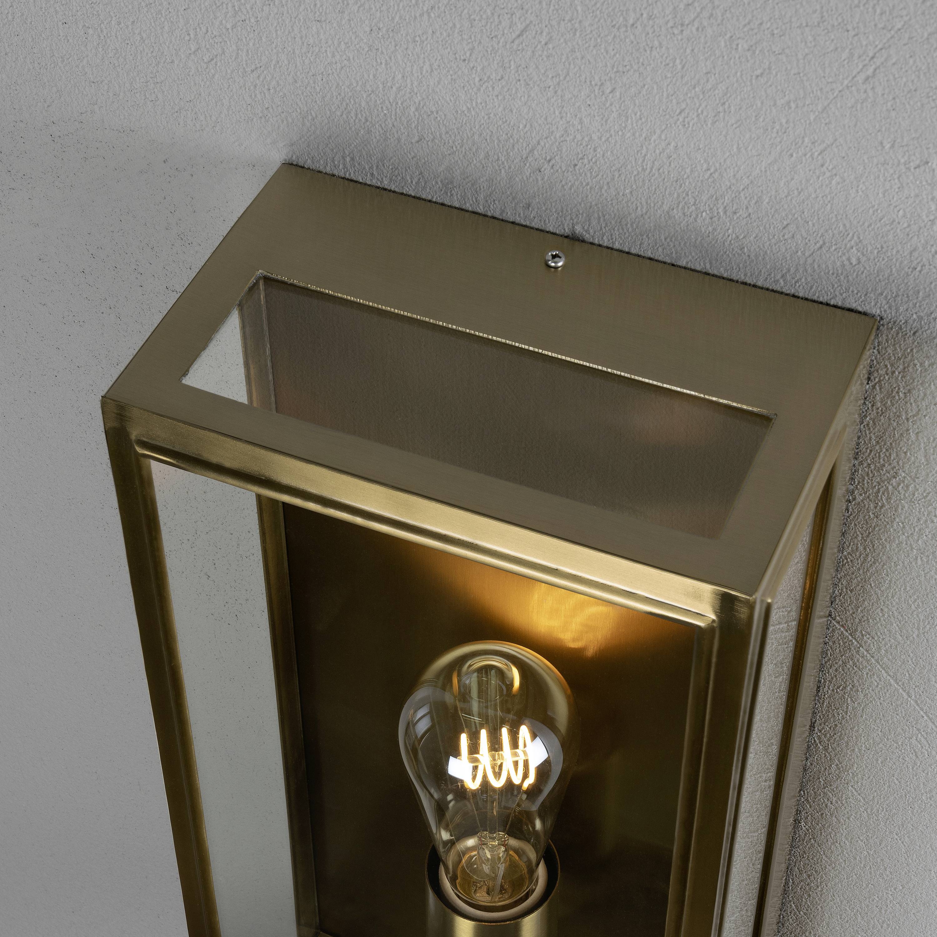 Kinkiet z kwadratową, złotą metalową ramką i widoczną żarówką, zamontowany na szarej ścianie.