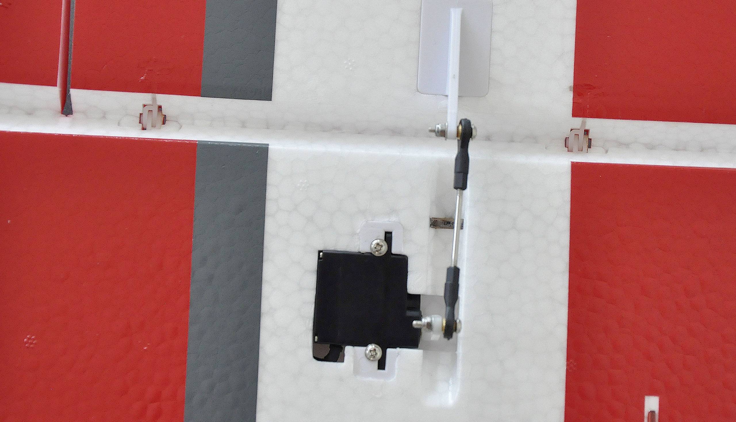 Zbliżenie skrzydła modelu samolotu z czerwono-białym podziałem kolorystycznym, mechanizmem dźwigniowym i serwosilnikiem sterującym.