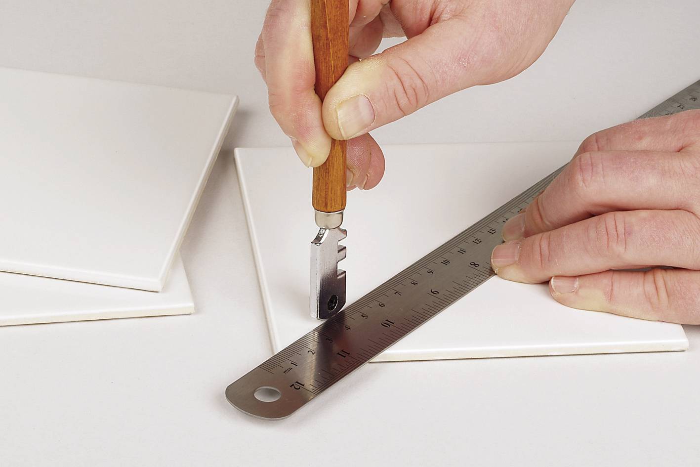 Osoba przycina białą płytkę za pomocą przecinaka do płytek i metalowej linijki.