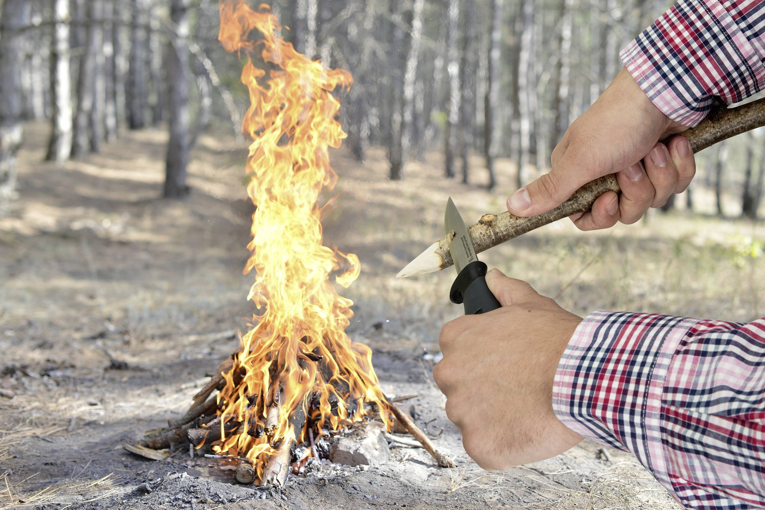 Osoba rzeźbi nożem kawałek drewna w lesie, podczas gdy w tle płonie małe ognisko.