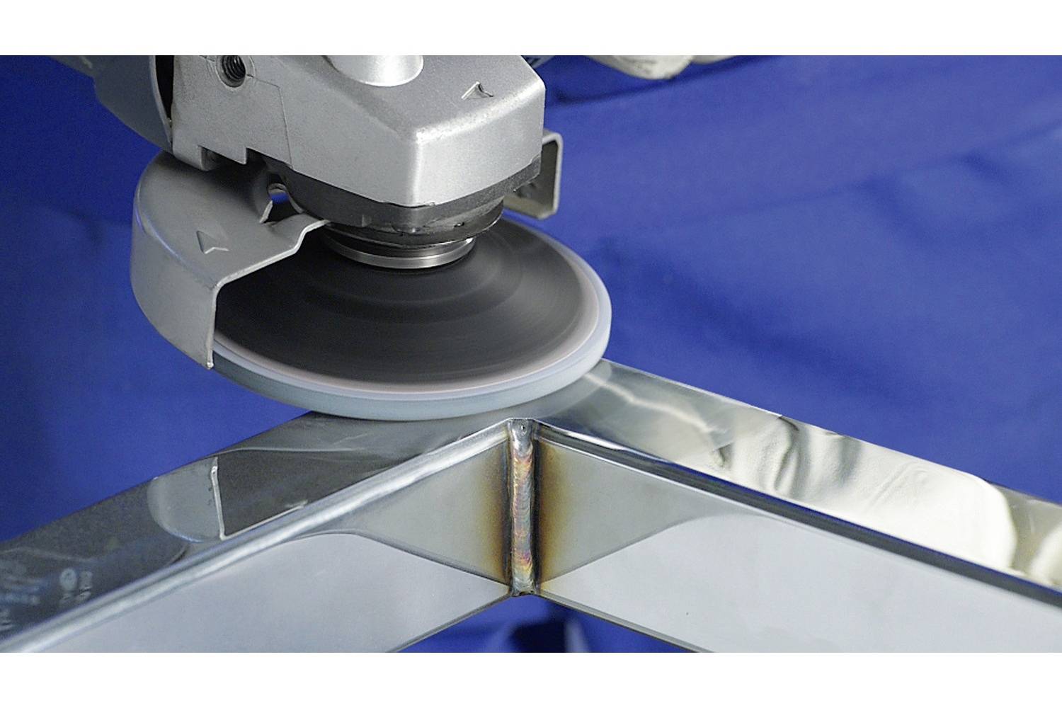 Szlifierka poleruje metalową krawędź. Błyszcząca powierzchnia odbija światło. Pracownik jest ubrany w niebieskie ubranie.