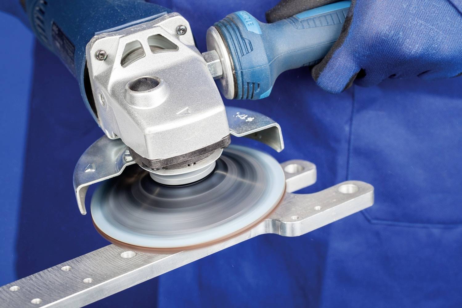 Osoba w niebieskich rękawiczkach szlifuje kawałek metalu szlifierką kątową. Narzędzie wytwarza iskry podczas szlifowania.