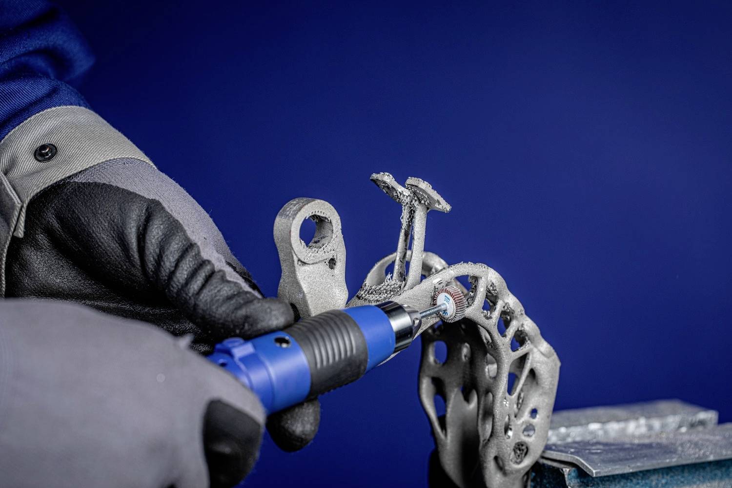 Osoba obrabia metalowy element za pomocą niebieskiego narzędzia, nosząc czarne i szare rękawice na niebieskim tle.