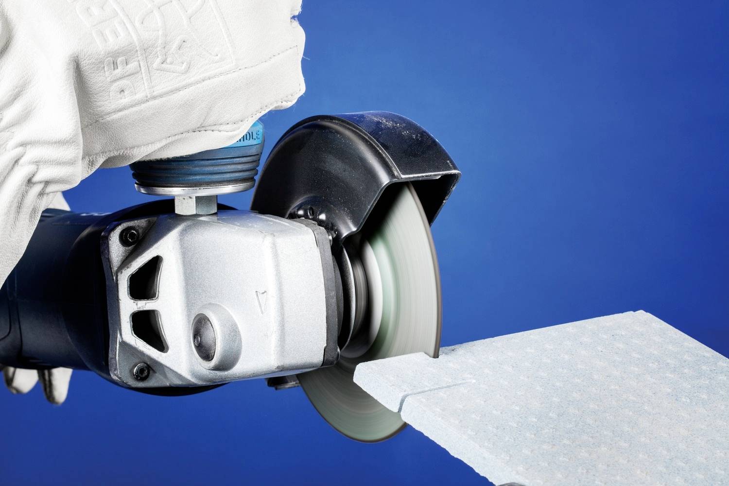 Szlifierka kątowa tnie białą płytę. Osoba nosi rękawice ochronne. Tło jest niebieskie.