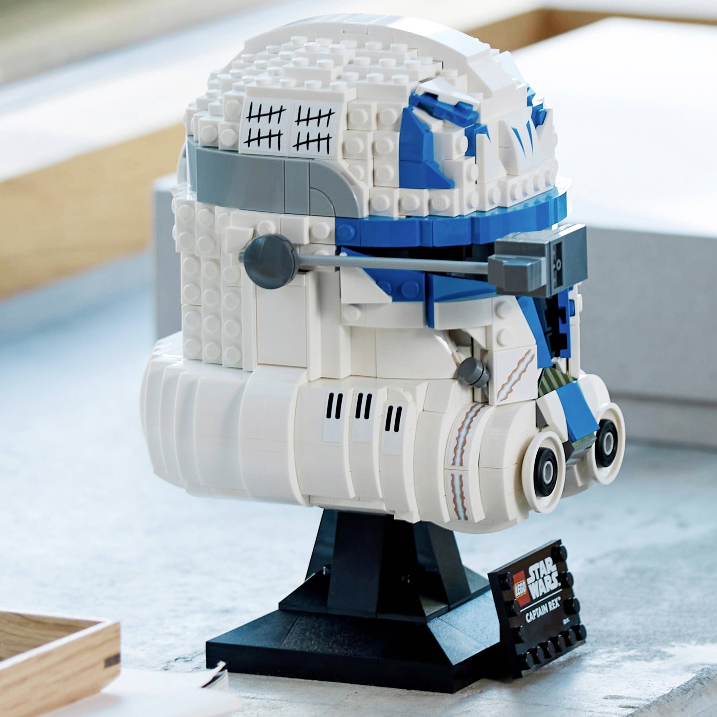 Model LEGO białego hełmu ze szczegółami w kolorze niebieskim, umieszczony na czarnym stojaku. Przypomina hełm z popularnego uniwersum science fiction.