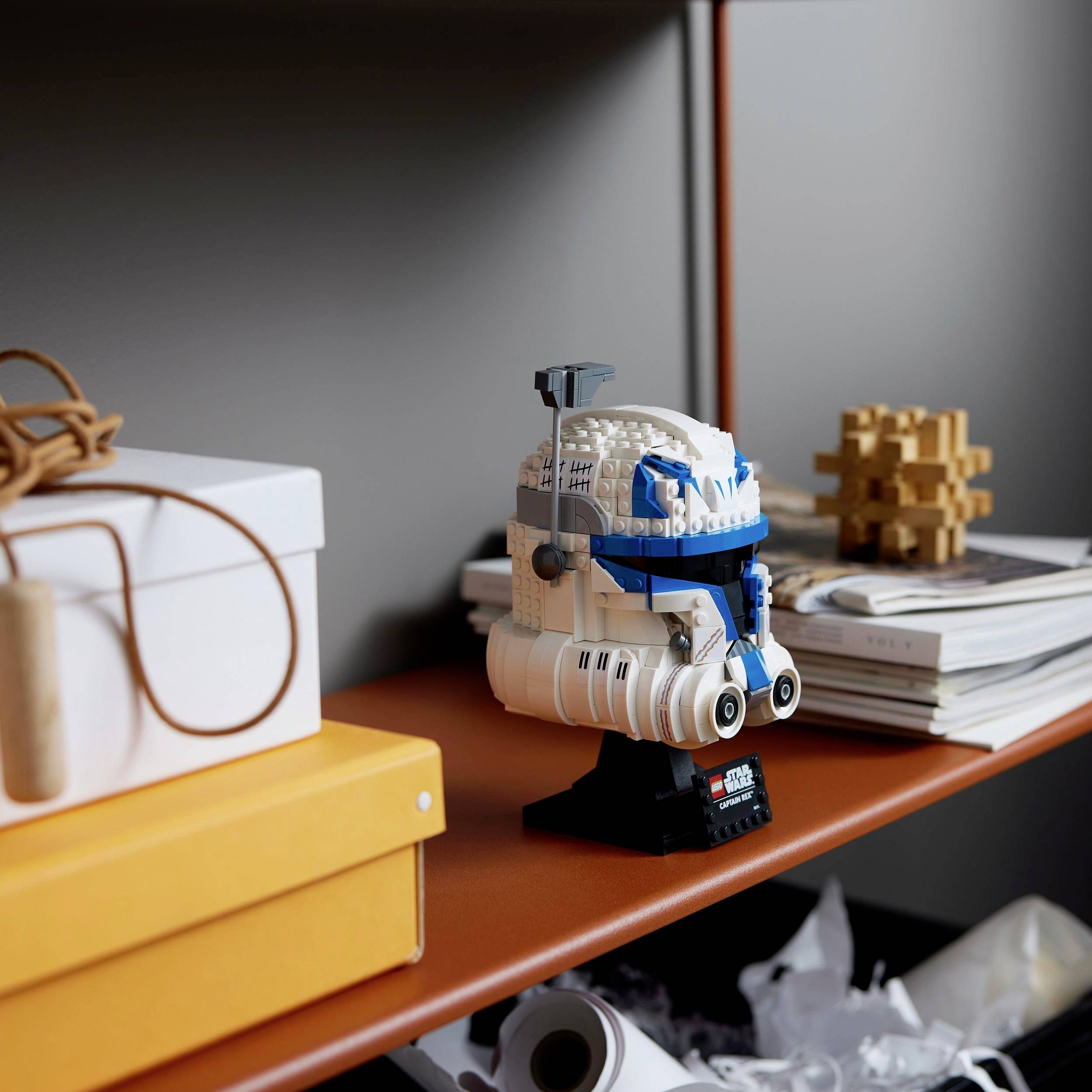 Robot Lego stoi na pomarańczowej półce, otoczony stosem książek i prezentem.