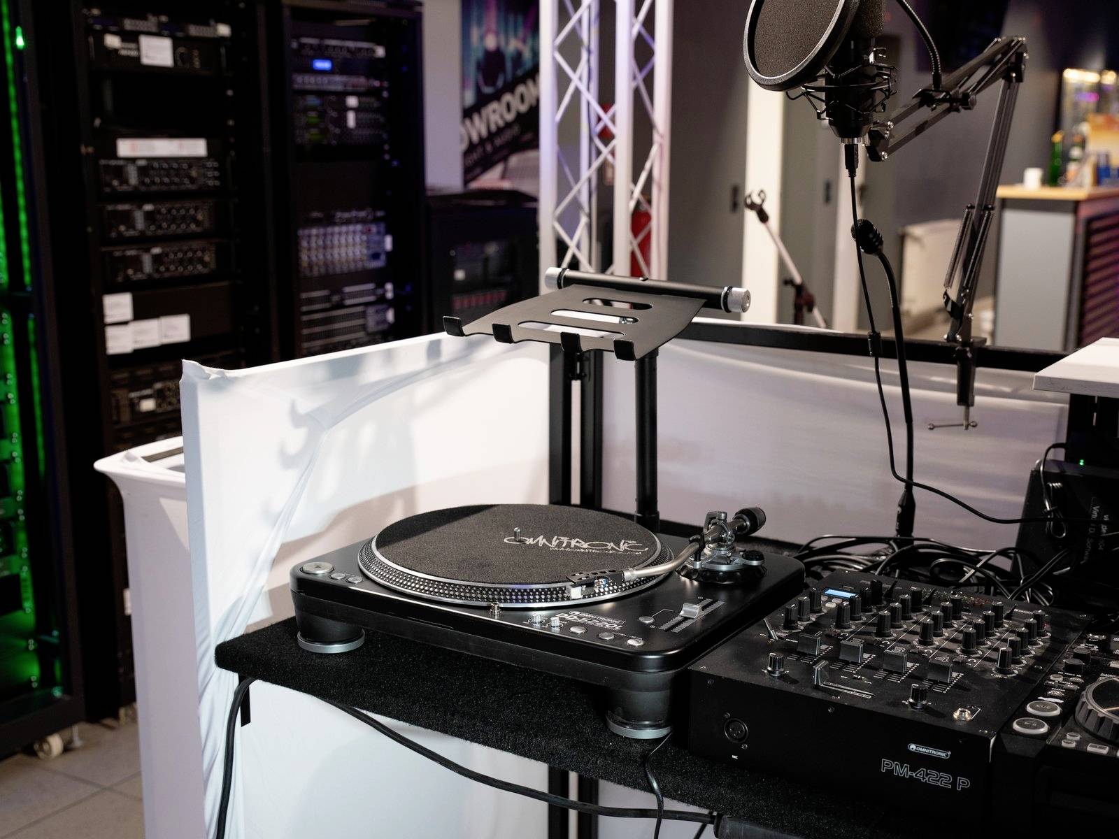 Studio nagrań z konsoletą mikserską, mikrofonem i gramofonem. W tle stojak z osprzętem technicznym i plakaty dekorujące pomieszczenie.