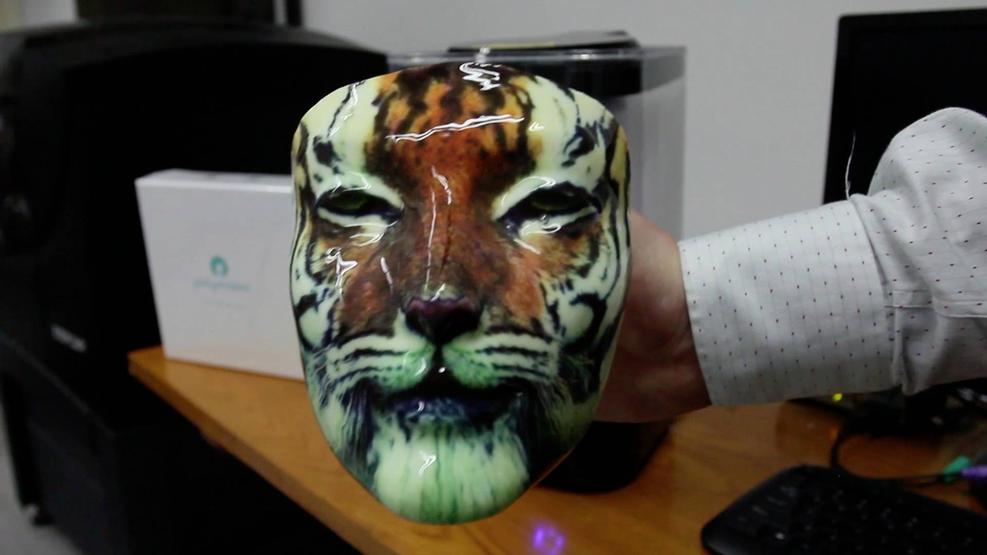 Ręka trzyma maskę przedstawiającą twarz tygrysa. Na tle widoczne są urządzenia biurowe.