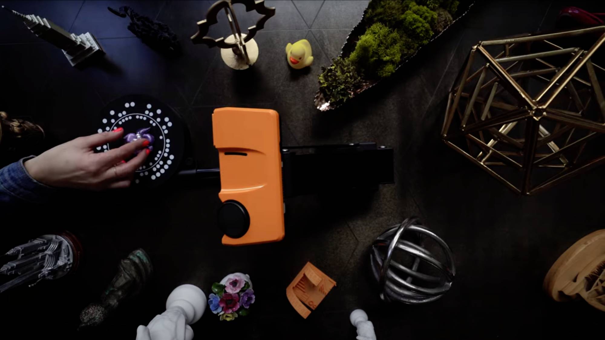 Osoba manipuluje pomarańczowym urządzeniem, otoczona dekoracyjnymi obiektami takimi jak figurki i geometryczne kształty na stole.