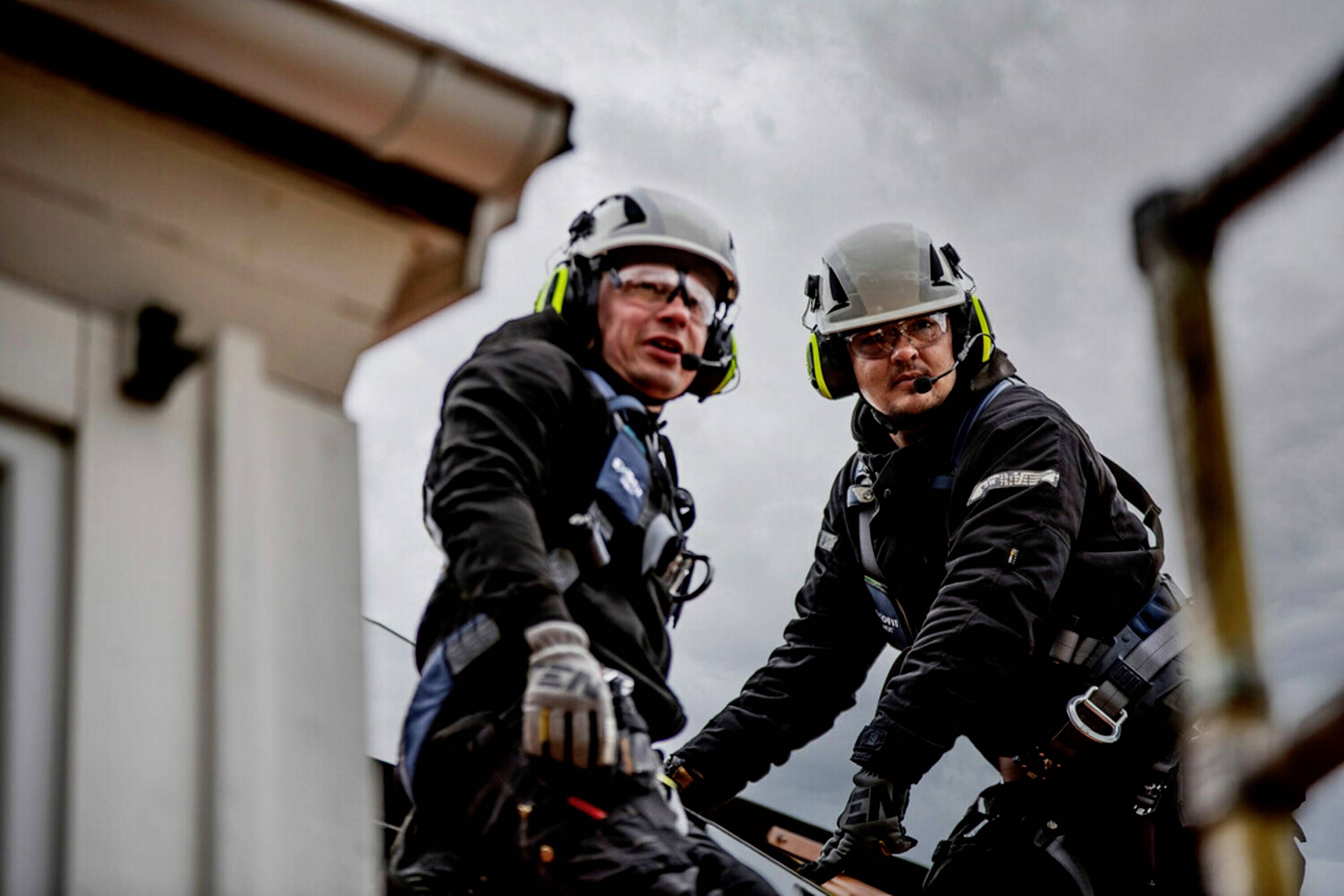 Dwie osoby w odzieży ochronnej i kaskach ochronnych stoją na dachu i patrzą w obiektyw, prawdopodobnie podczas prac konserwacyjnych.