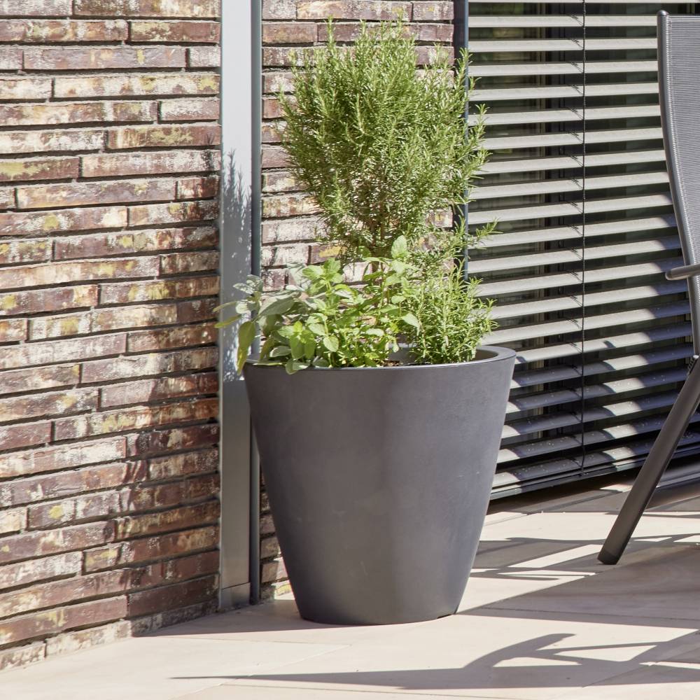 Duży czarny doniczka stoi na tarasie z ceglaną ścianą i żaluzjami w tle. Rosną w niej rozmaryn i bazylia.