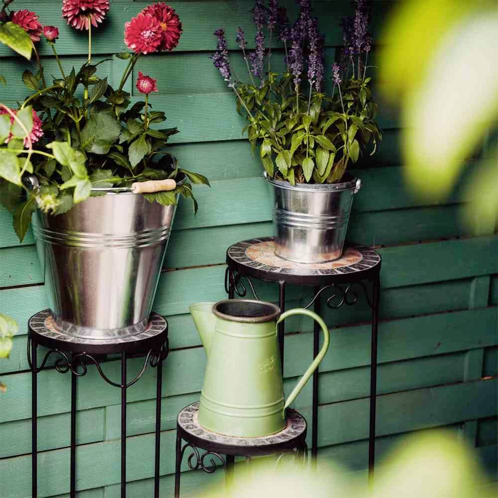 Dwa wiadra z różowymi i fioletowymi kwiatami stoją na półkach przed zieloną drewnianą ścianą, obok znajduje się zielona konewka.